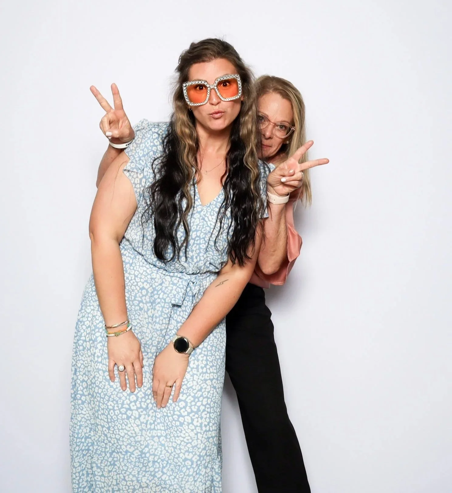 Two women making peace signs and wearing fun accessories, posing against a white background.