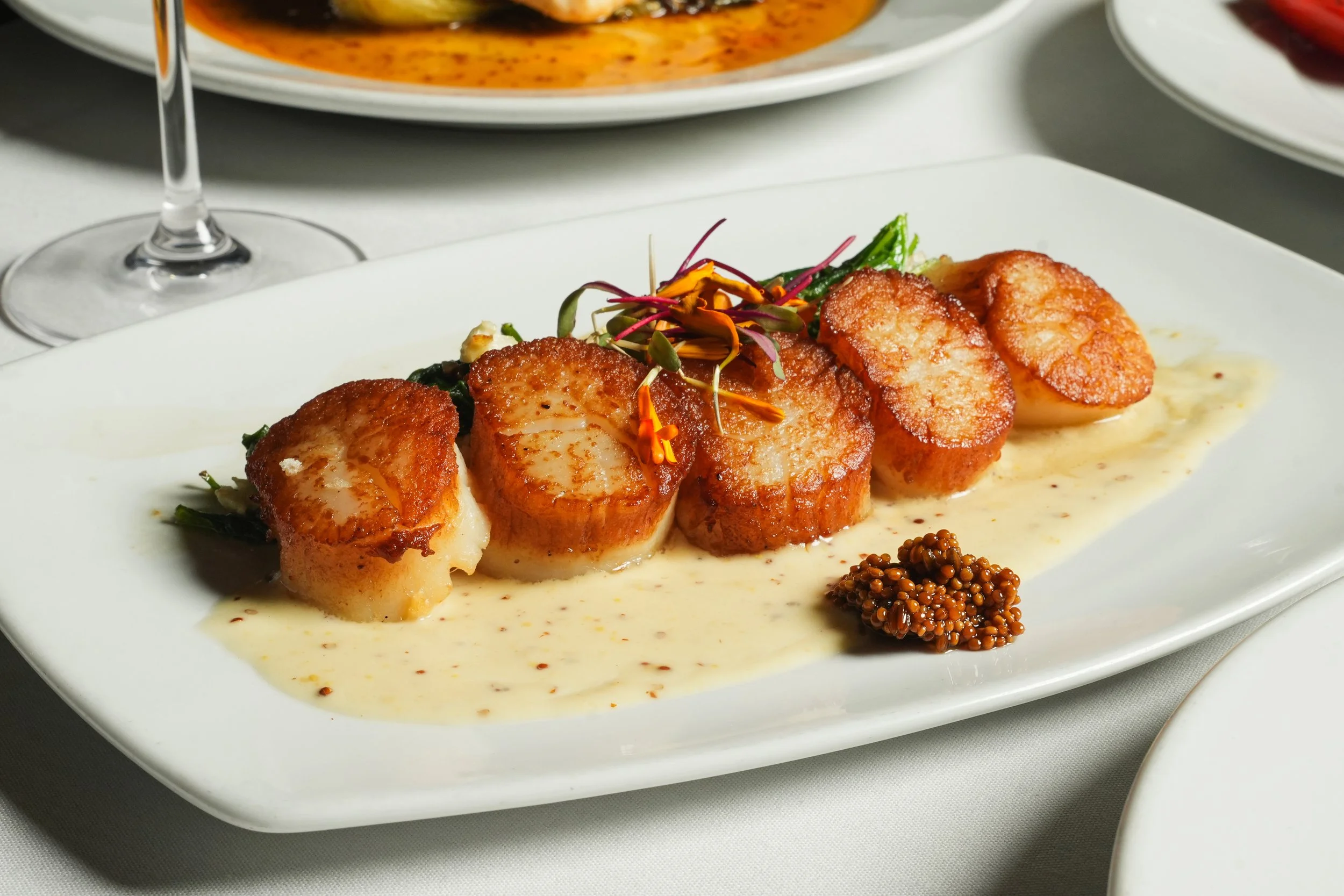 Seared scallops served on a white rectangular plate with a creamy sauce, garnished with microgreens and mustard seeds, with a bowl of red sauce in the background.