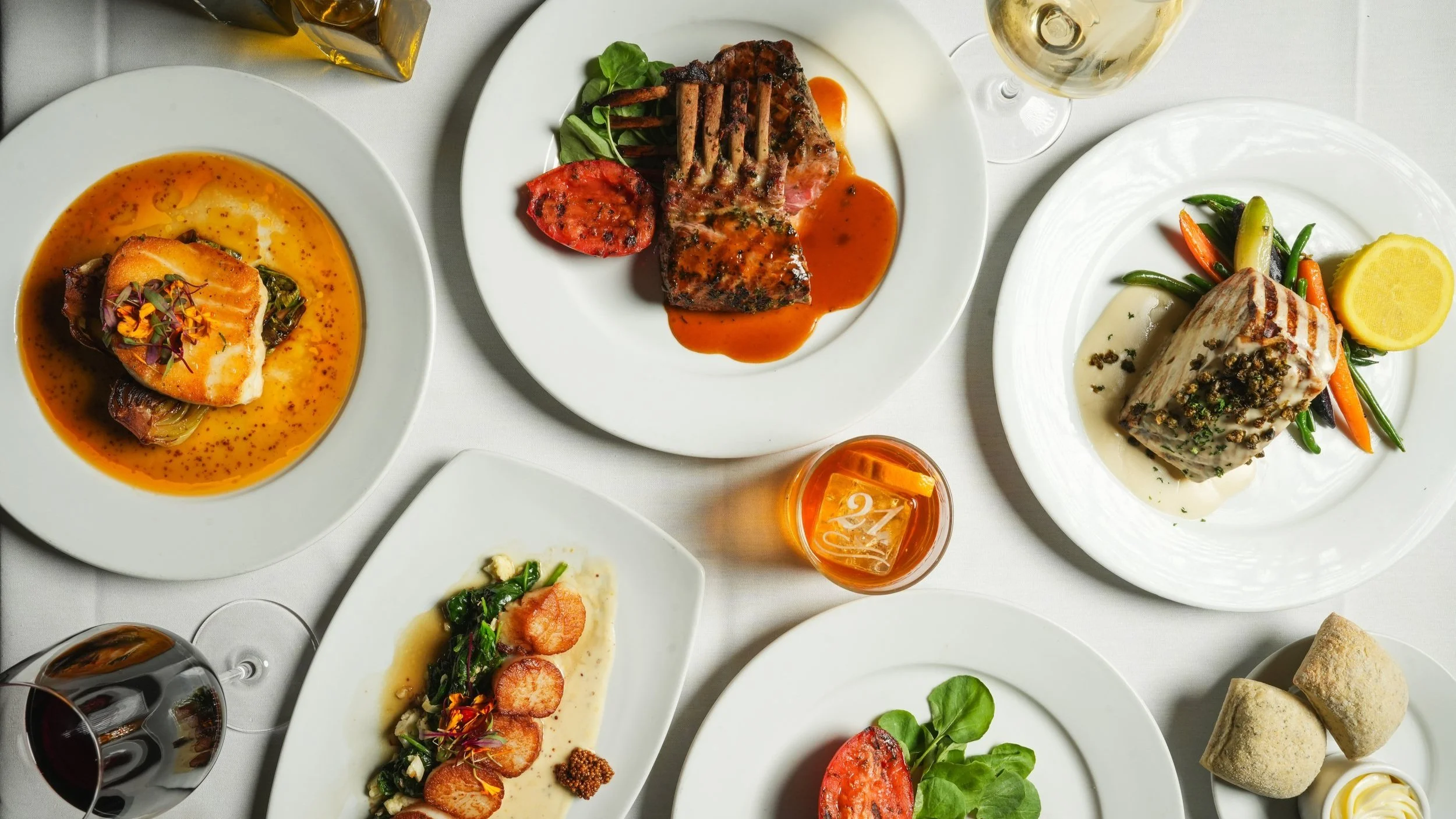 Assorted gourmet dishes on white plates, including fish, lamb, beef, vegetables, bread rolls, and glasses of wine and a cocktail, all arranged on a white tablecloth.