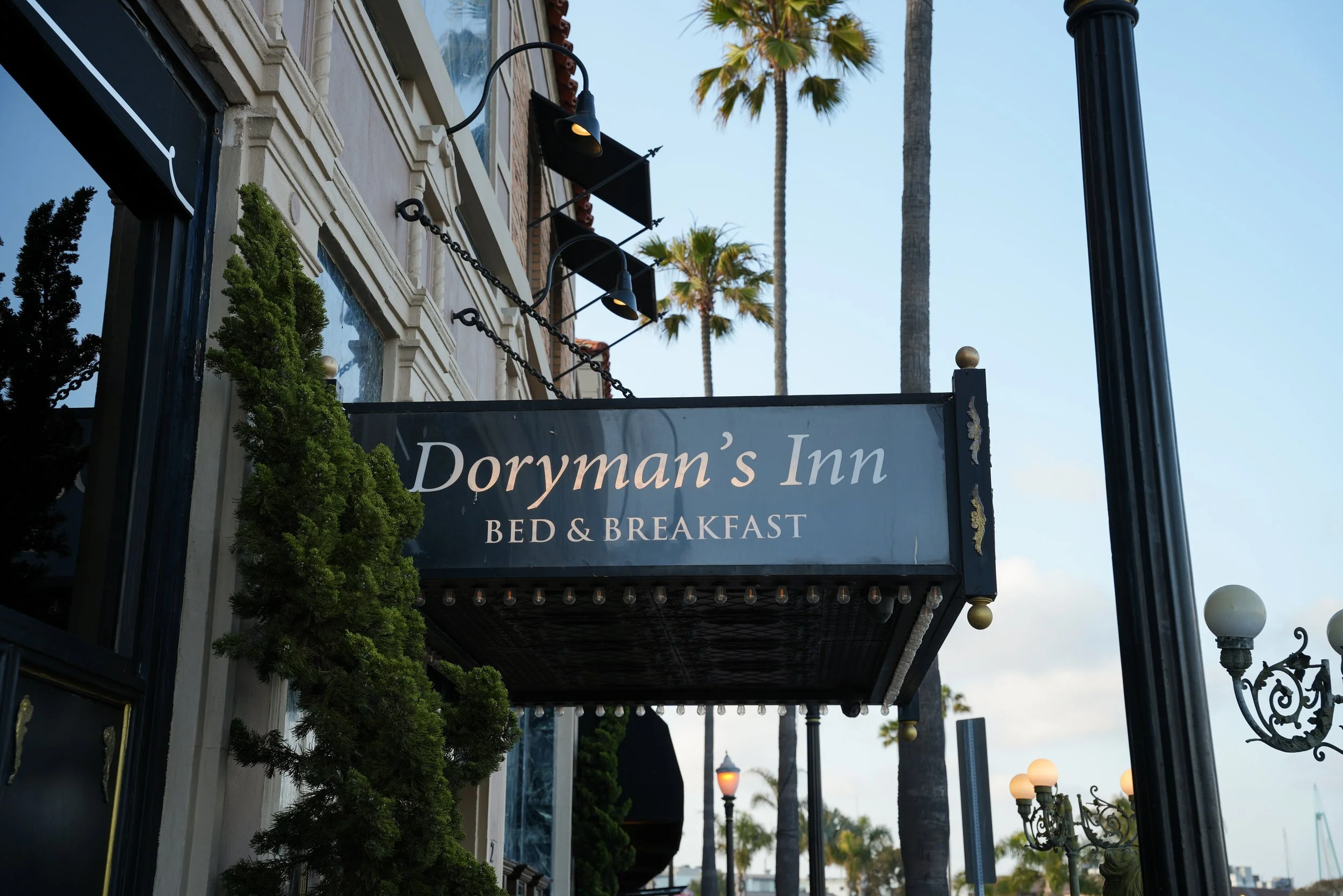 Sign reading 'Doryman's Inn Bed & Breakfast' hanging outside a building with decorative lighting, palm trees, and vintage-style street lamps in the background.