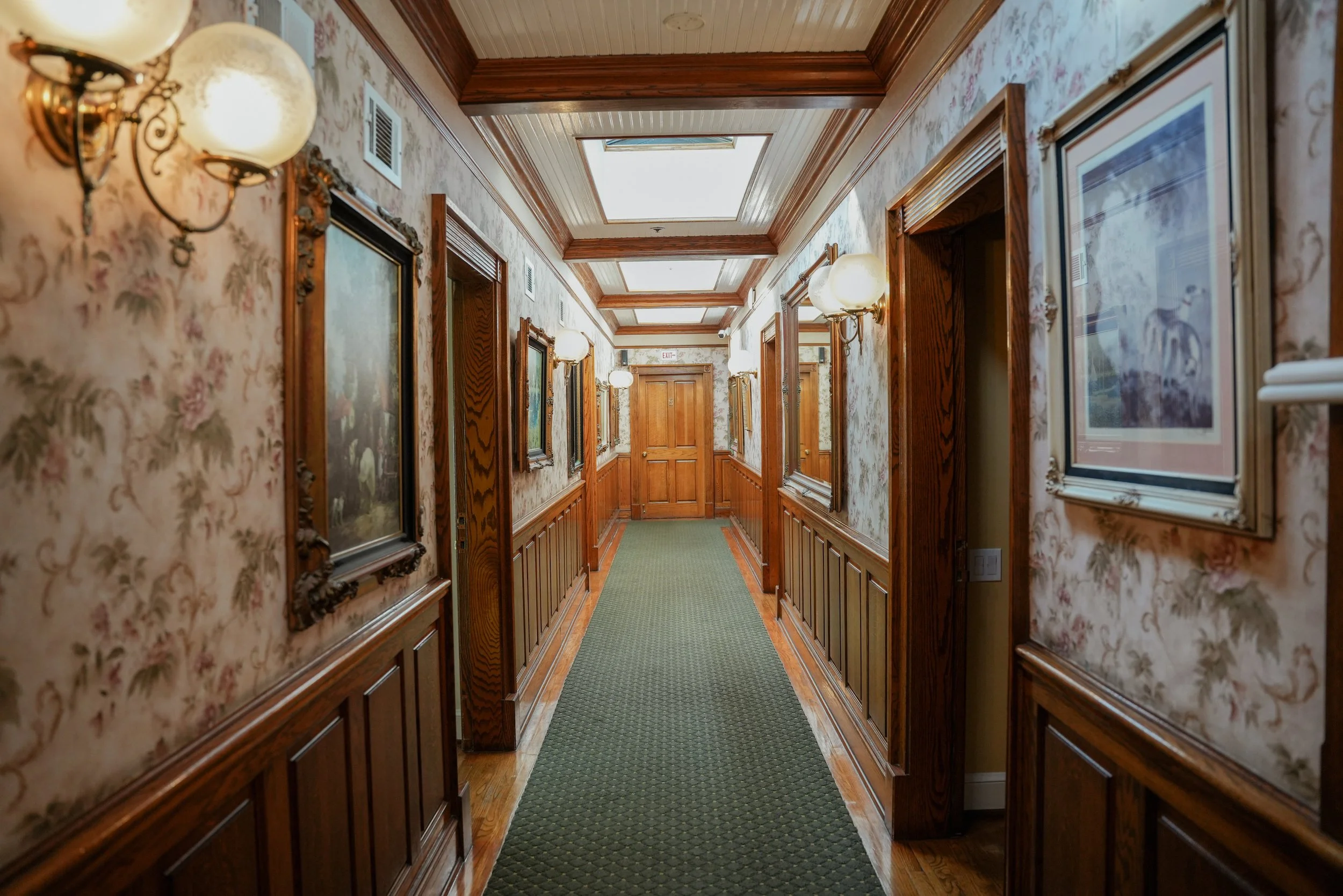 A hallway with floral wallpaper, wooden wainscoting, framed paintings, wall-mounted lamps, and skylights overhead.