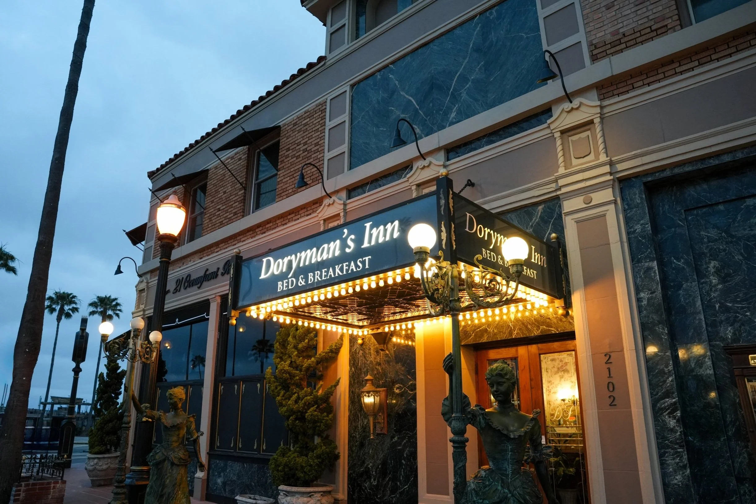 Exterior of Doryman's Inn bed and breakfast during evening, with a black signboard illuminated by lights, decorative statues of women outside, and palm trees in the background.