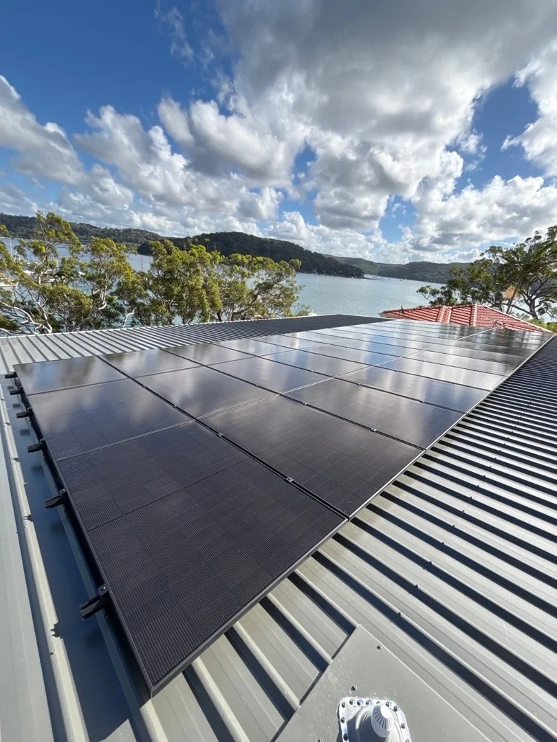 View of solar panels installed on a corrugated metal roof with a lake, trees, and cloudy sky in the background.