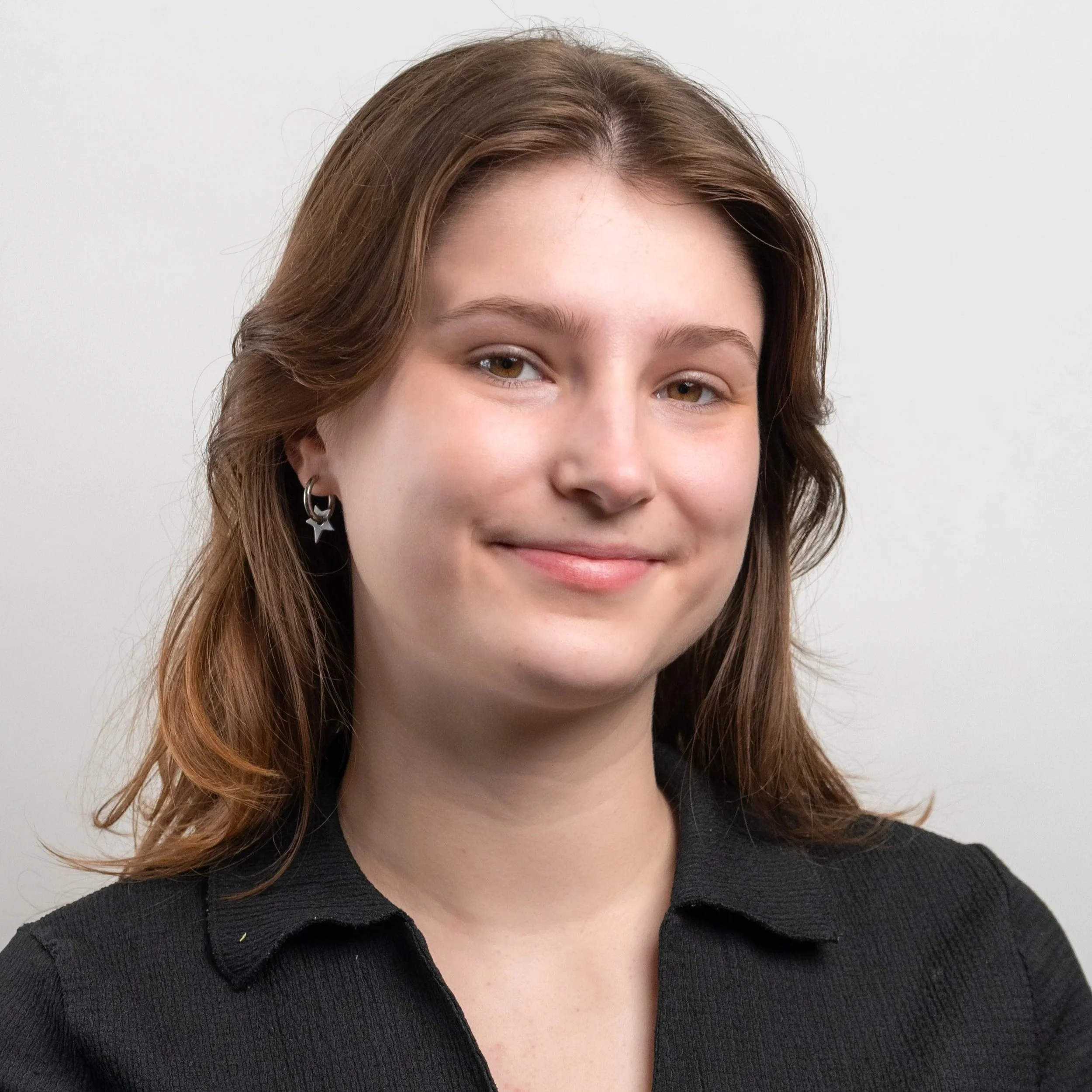 A young woman with long brown hair smiling outdoors in front of fall foliage, wearing a blue and white checkered dress and a beaded necklace.