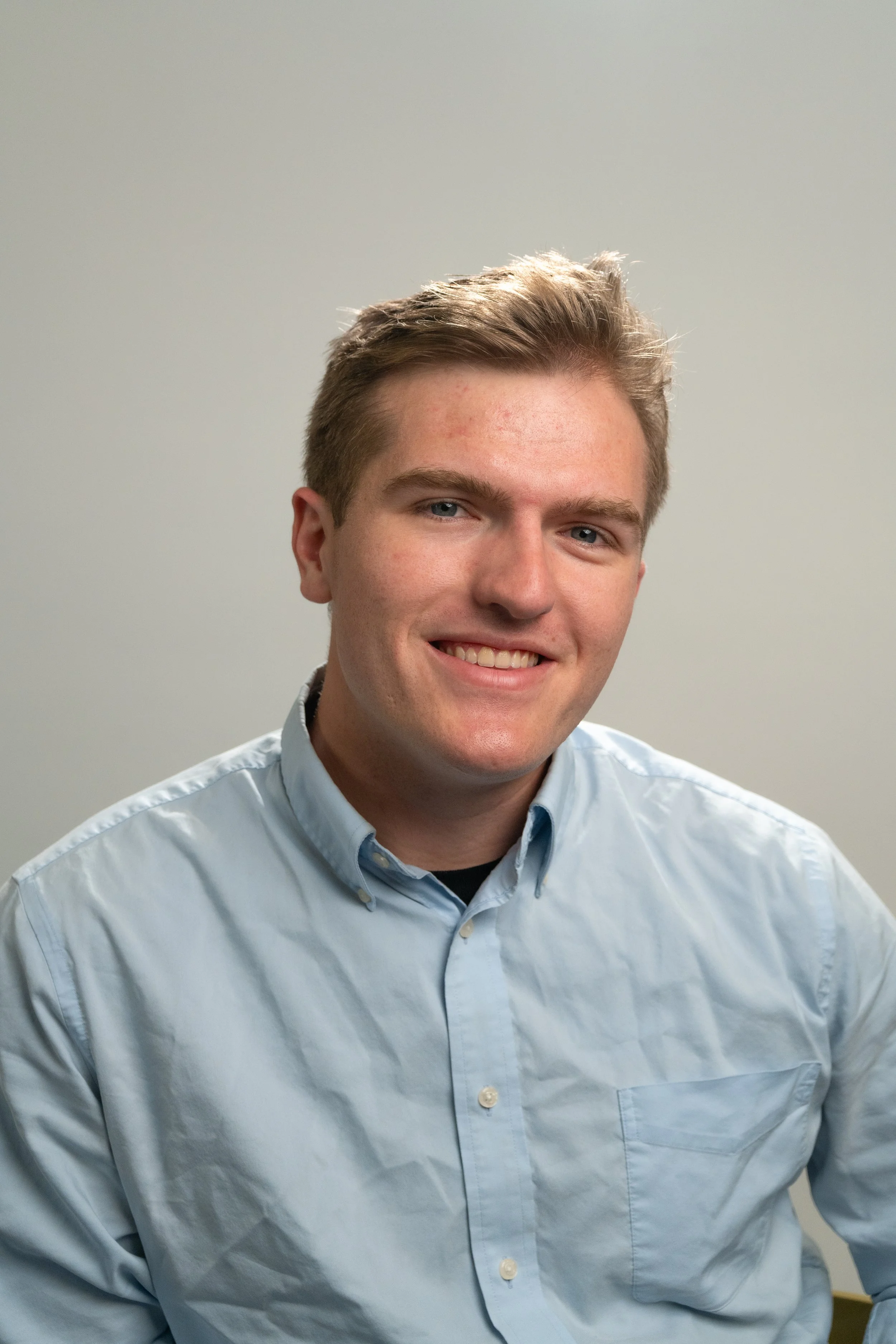A man with short, light brown hair and a beard, wearing a blue button-up shirt, smiling and sitting at a table with a plant and a framed picture in the background.