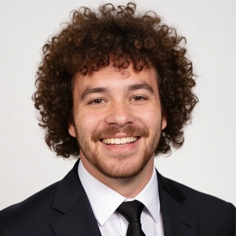 A smiling man with curly hair and a beard, wearing a black jacket and a chain necklace, in a dimly lit setting with string lights in the background.