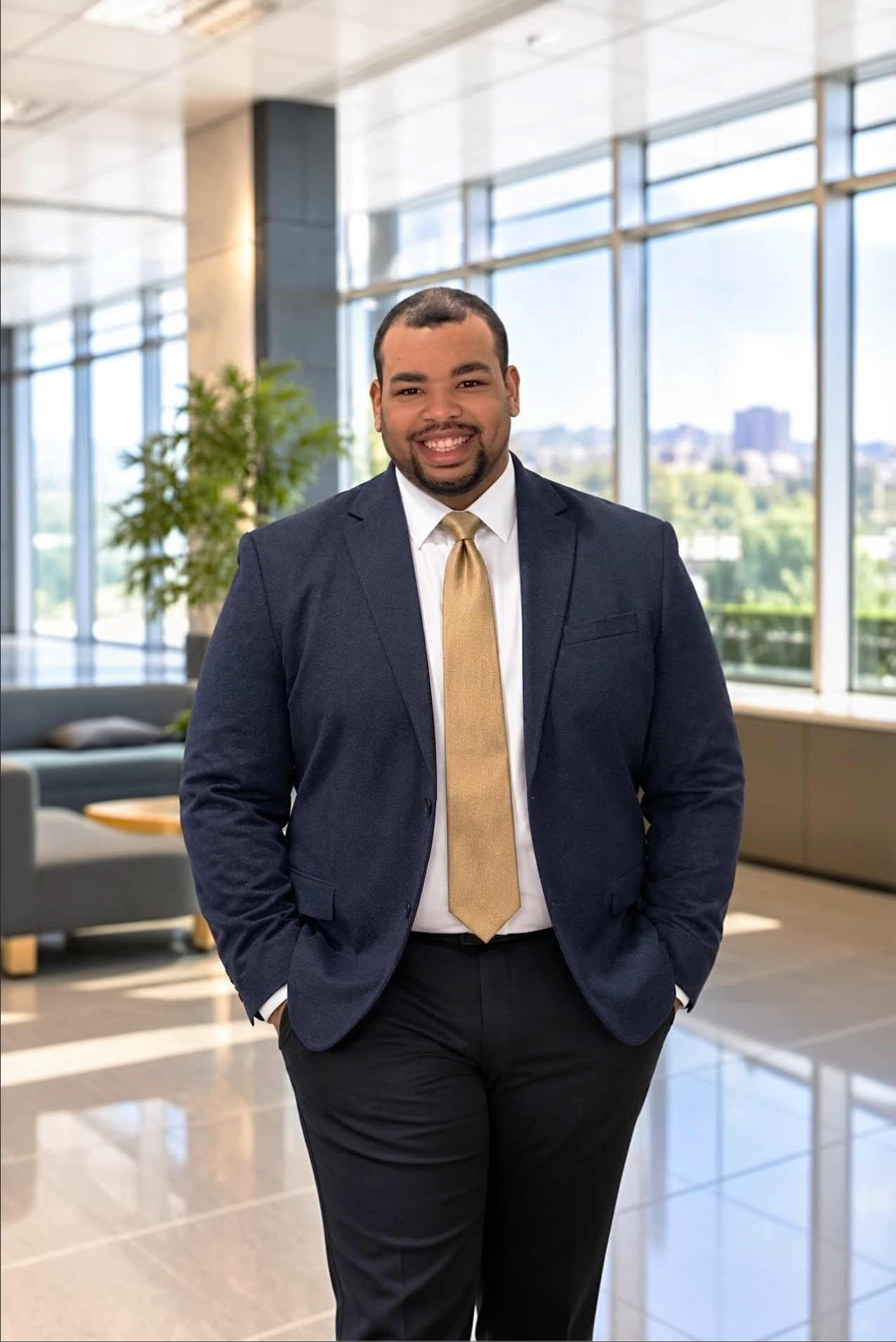 A smiling man in a navy suit, white shirt, and gold tie standing in an office lobby with large windows and greenery outside.