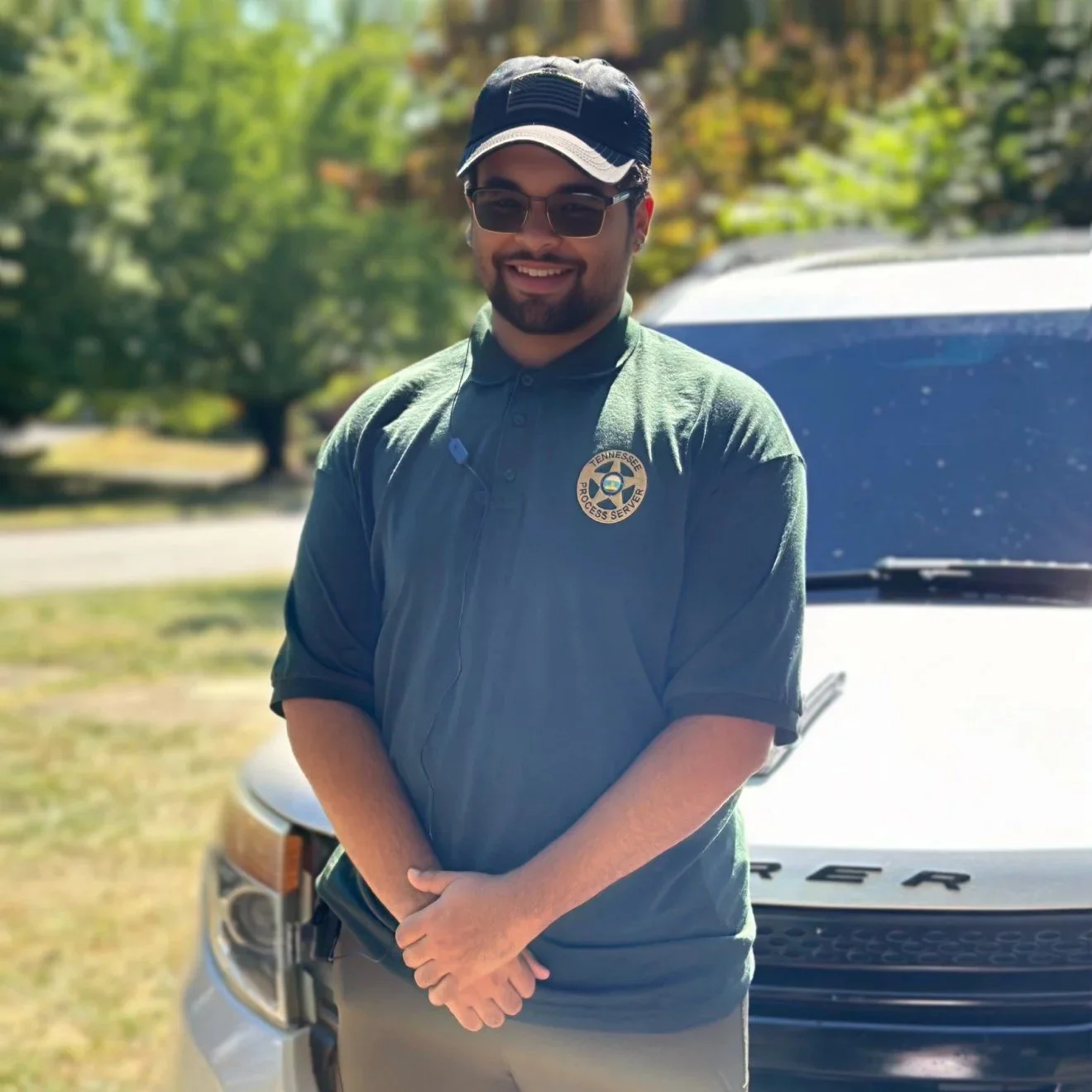 A man smiling, wearing sunglasses, a hat, and a dark green shirt with a Tennesse Process Server badge, standing outdoors next to a white vehicle with a tree-lined background.