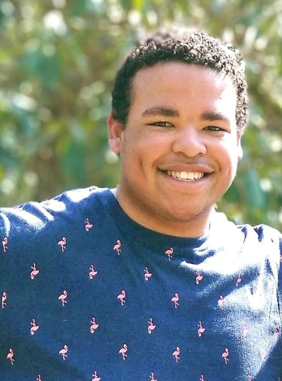 A person smiling outdoors with greenery in the background, wearing a navy blue shirt with pink flamingo patterns.