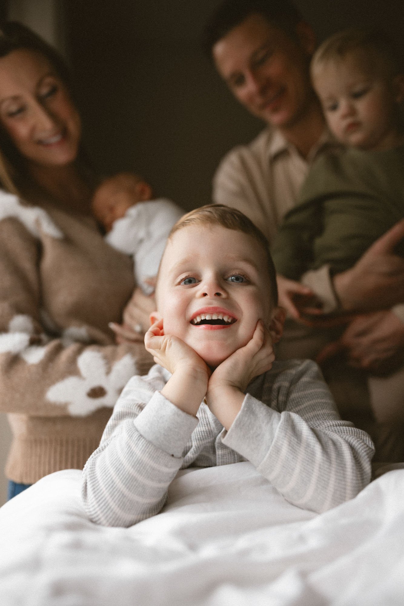 A smiling toddler poses on a bed during a cozy in-home newborn family session, with parents holding a newborn and another child in a softly lit bedroom.