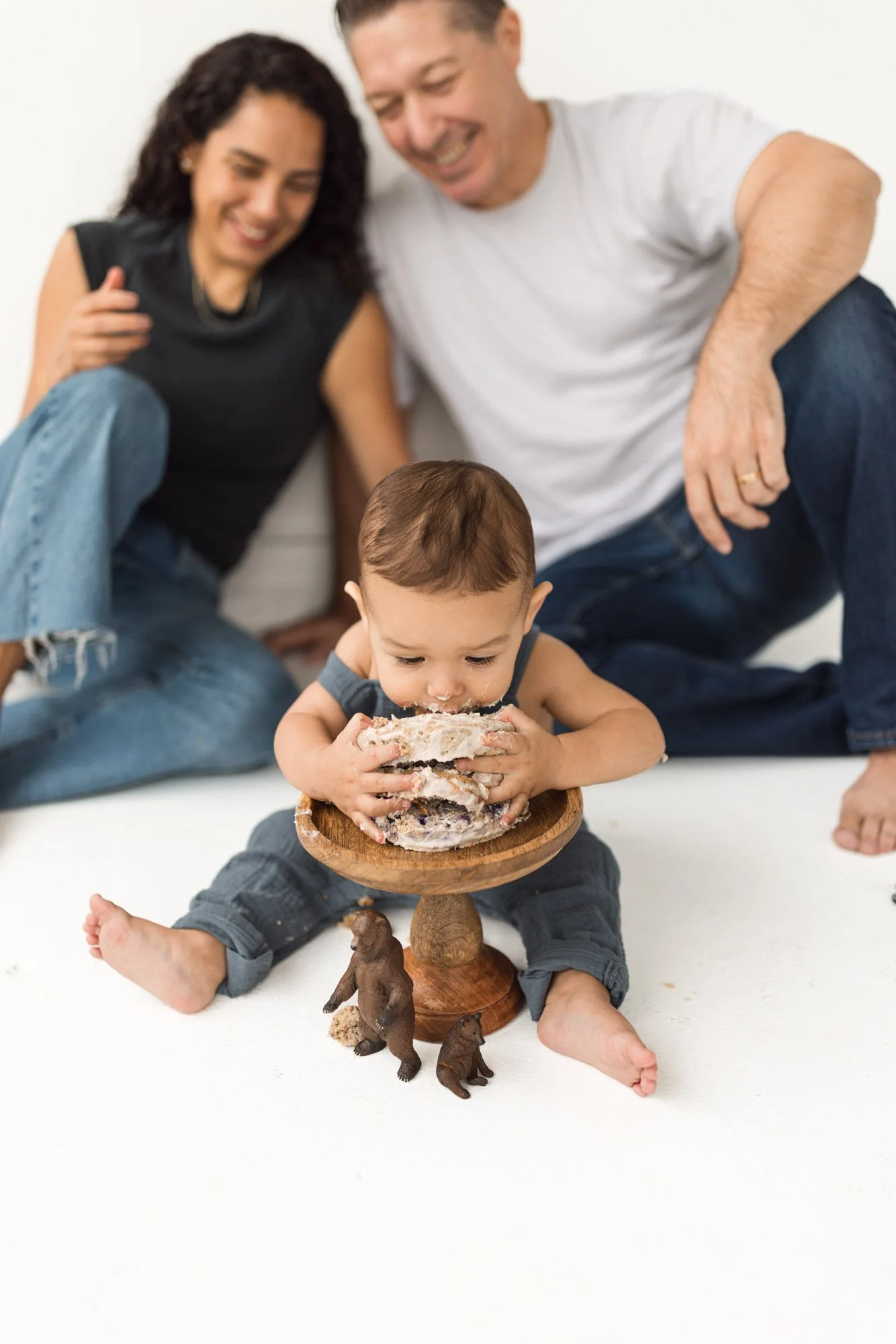 One-year milestone cake smash session with baby enjoying cake while parents smile in the background, captured by a Seattle family photographer. Natural, candid family interaction in a bright studio setting with neutral tones and timeless styling.