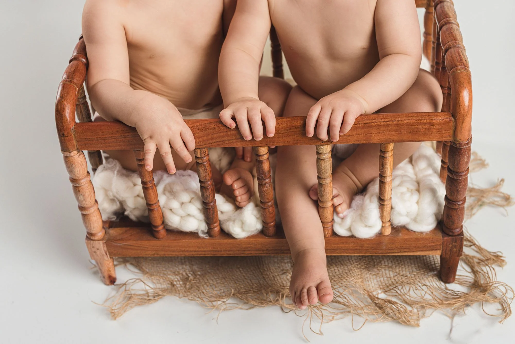 Twin babies sitting closely together on a wooden bench, showing tiny hands and feet during a milestone photography session for a first birthday in Gig Harbor, Washington