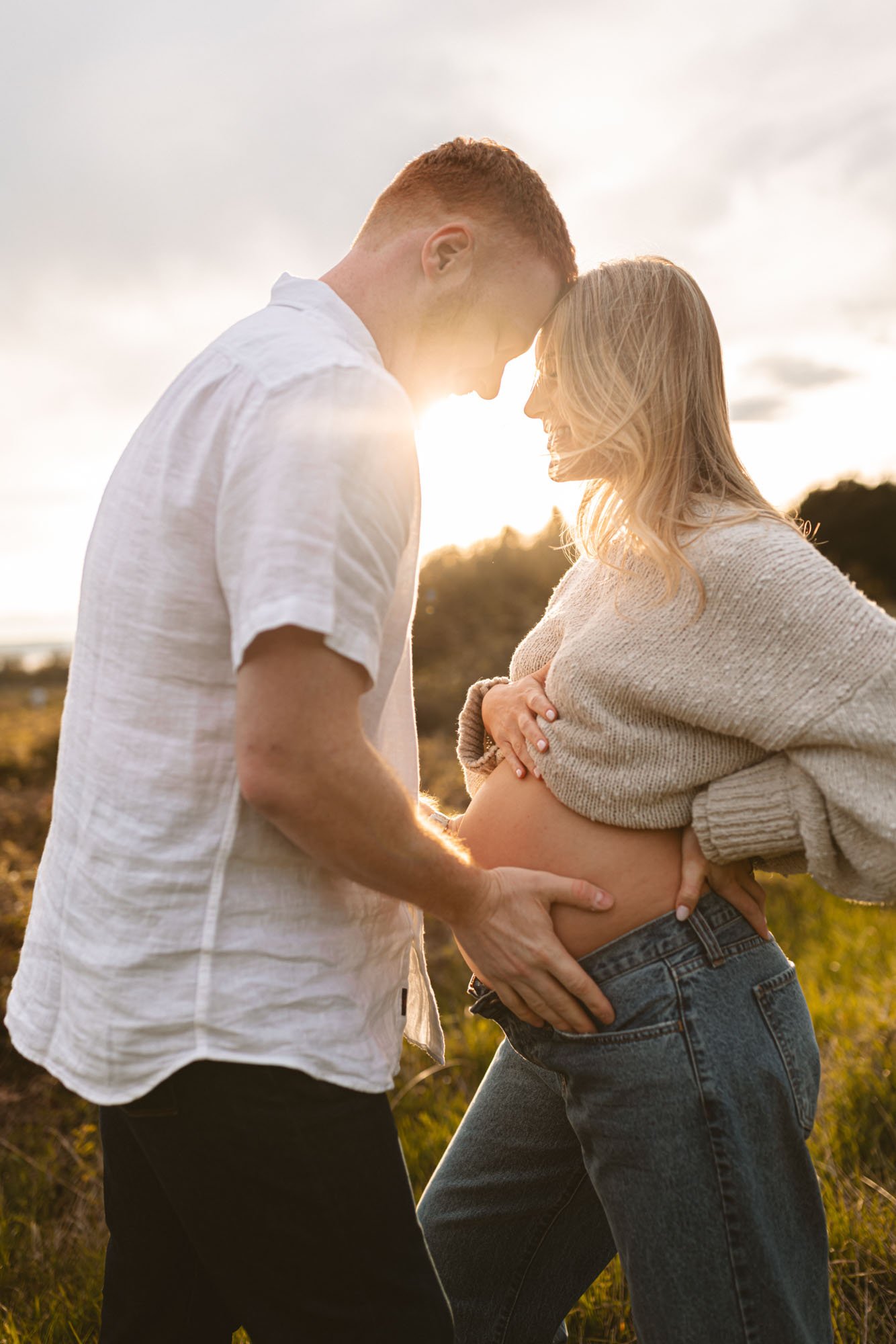 Expecting couple leaning into each other in warm golden light at Discovery Park, Seattle, capturing a soft, intimate moment as they connect with their baby.