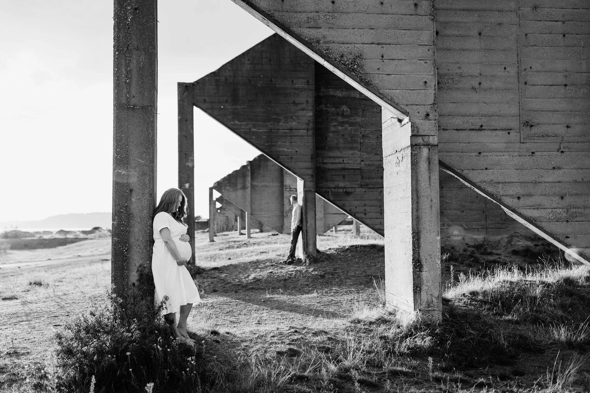 Black and white maternity portrait of a mother standing in an open landscape with architectural lines, creating a timeless and artistic feel in Tacoma