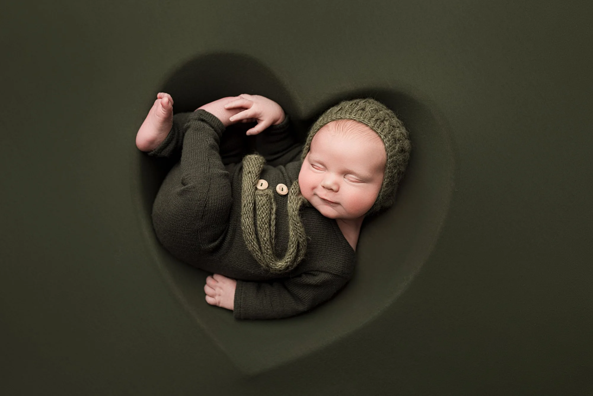 Sleeping newborn baby boy wearing a dark green outfit and knit bonnet during a Seattle studio newborn session.