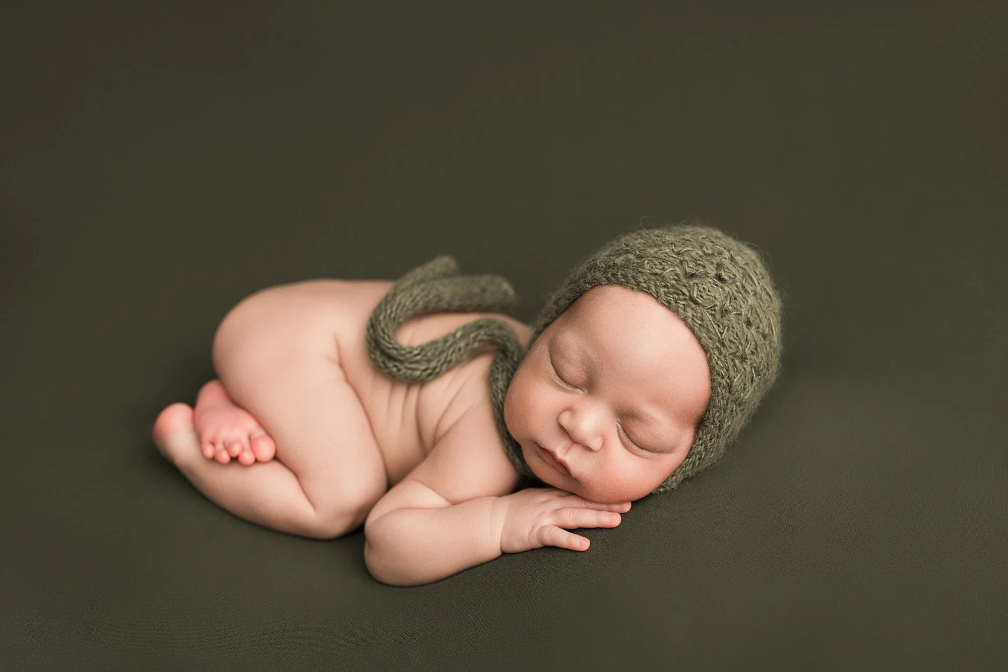 Sleeping newborn baby wearing a knitted green bonnet on a coordinating backdrop