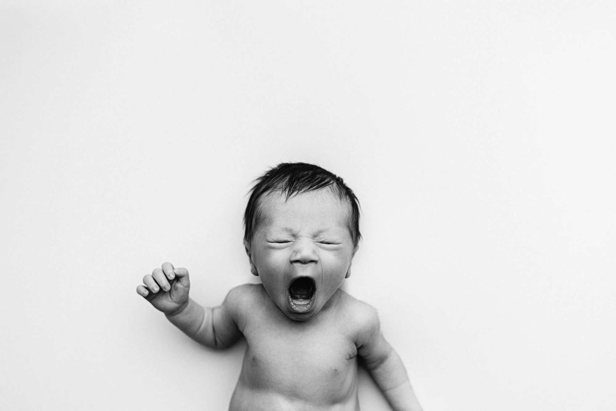Black and white photo of a newborn baby yawning with eyes closed and one arm raised.