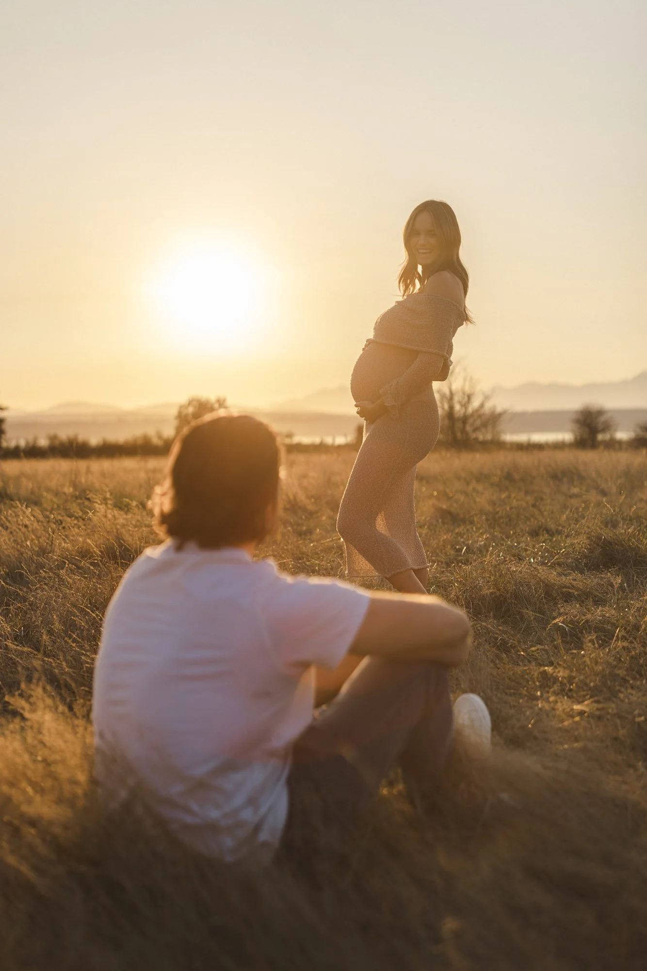 Seattle maternity photography session at sunset with expecting mother in a field and partner sitting nearby, golden hour pregnancy portrait