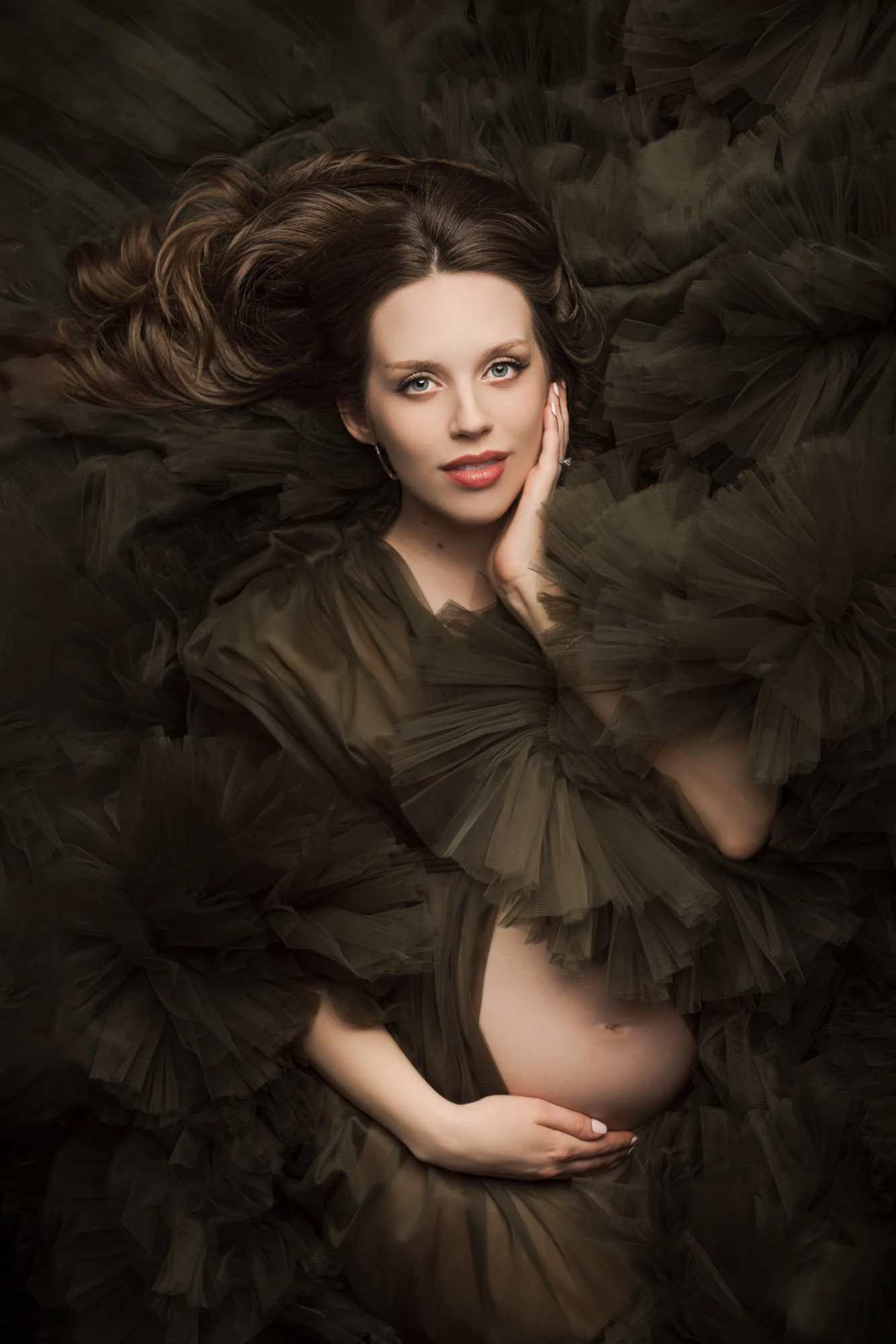 Fine art maternity portrait of an expecting mother in an elegant gown, photographed in a professional studio with dramatic lighting.