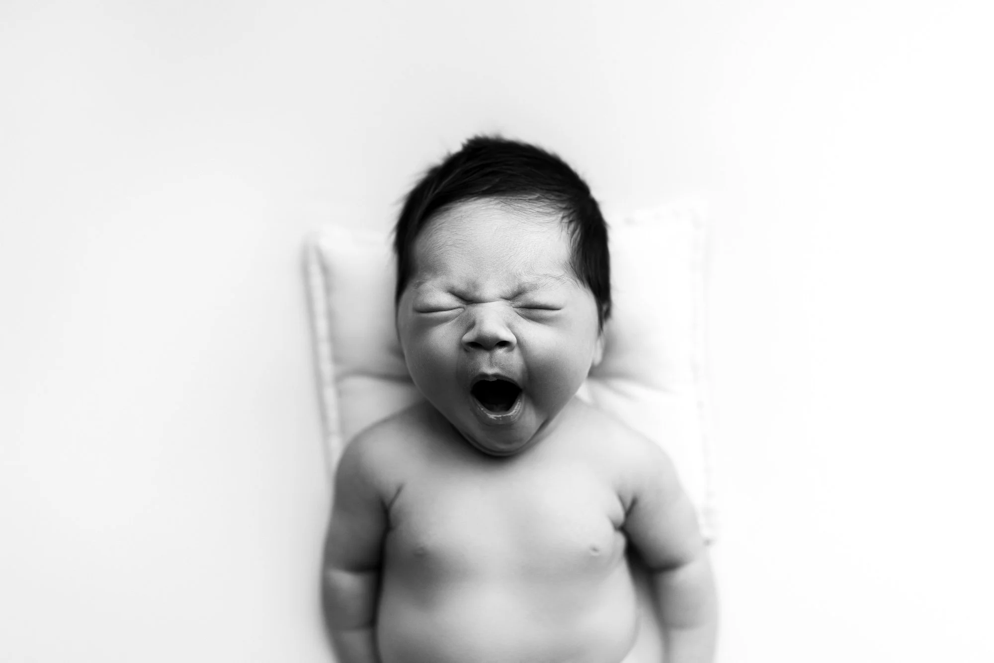 A black and white photo of a baby with dark hair yawning while lying on a small pillow against a plain background.
