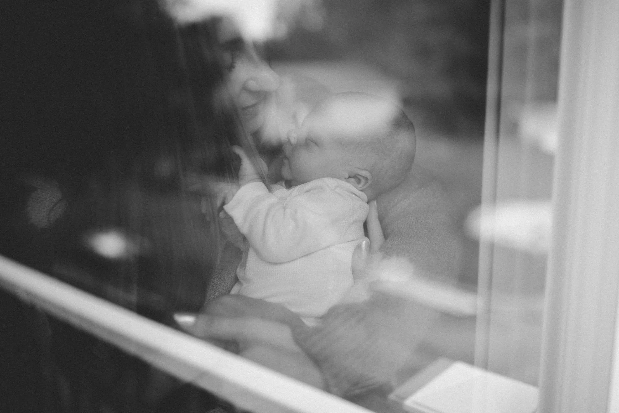 A mother holds her newborn by a window, seen through a soft reflection, during a calm in-home newborn photography session in Seattle.