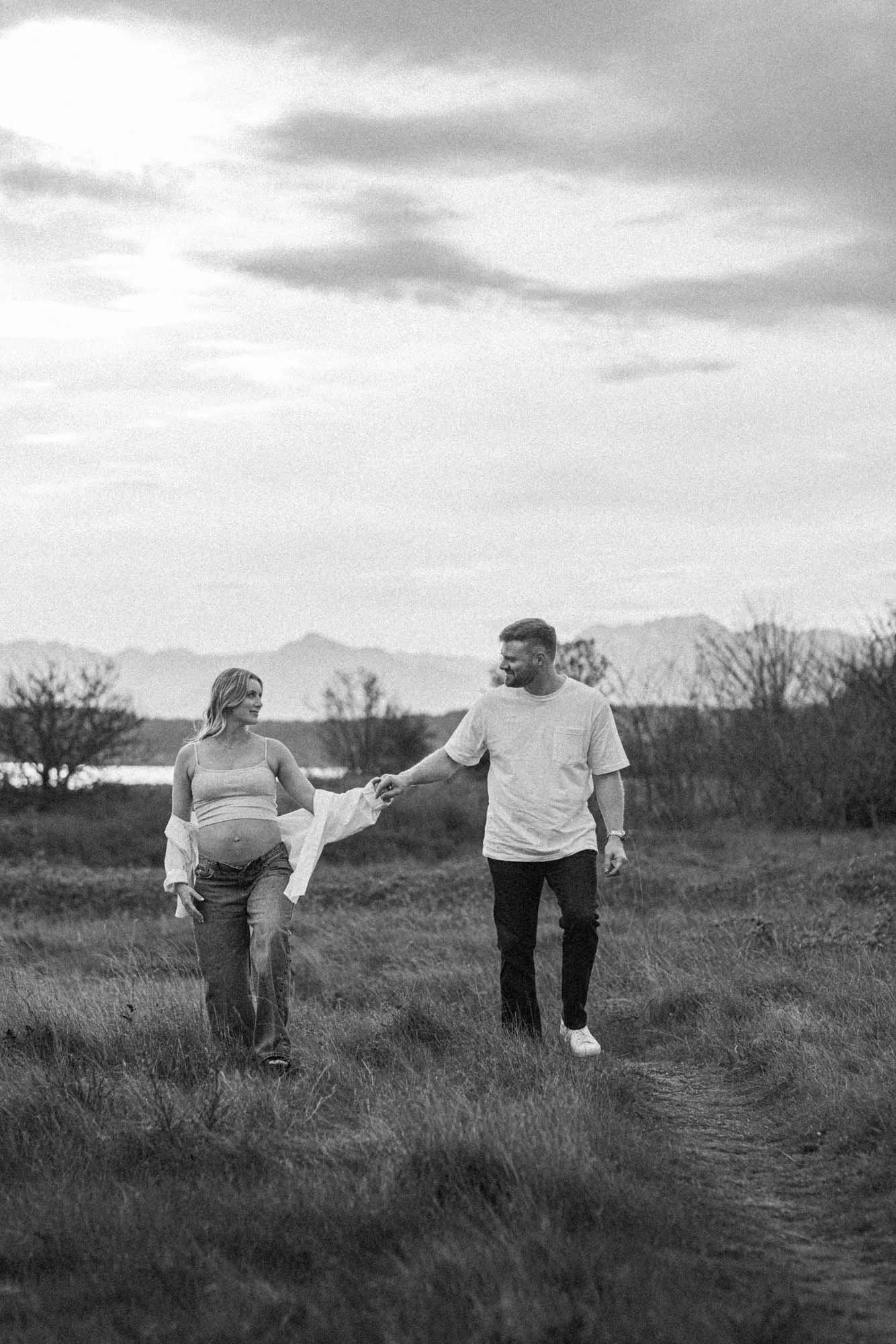 Couple walking hand in hand through the open field at Discovery Park in Seattle, capturing a relaxed and connected maternity moment with soft, moody tones.