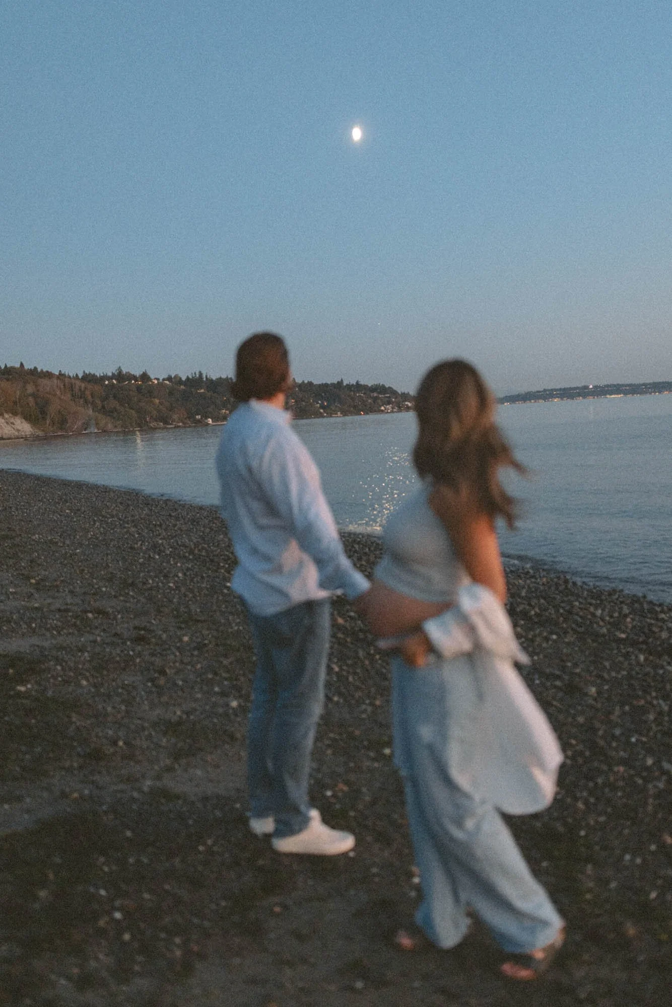 Expecting couple holding hands by the water at dusk, looking toward the moon during a romantic maternity session in Seattle
