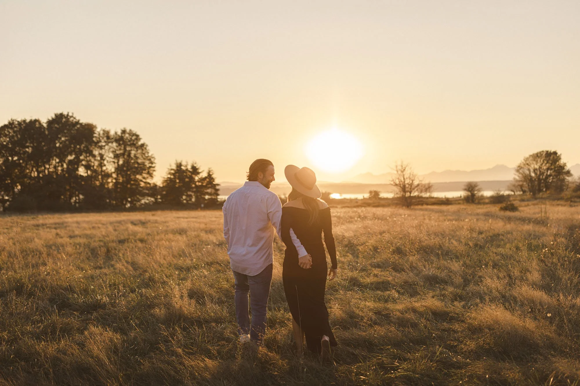 Expecting couple walking hand in hand through a sunlit field at sunset during a warm and natural maternity session in Seattle