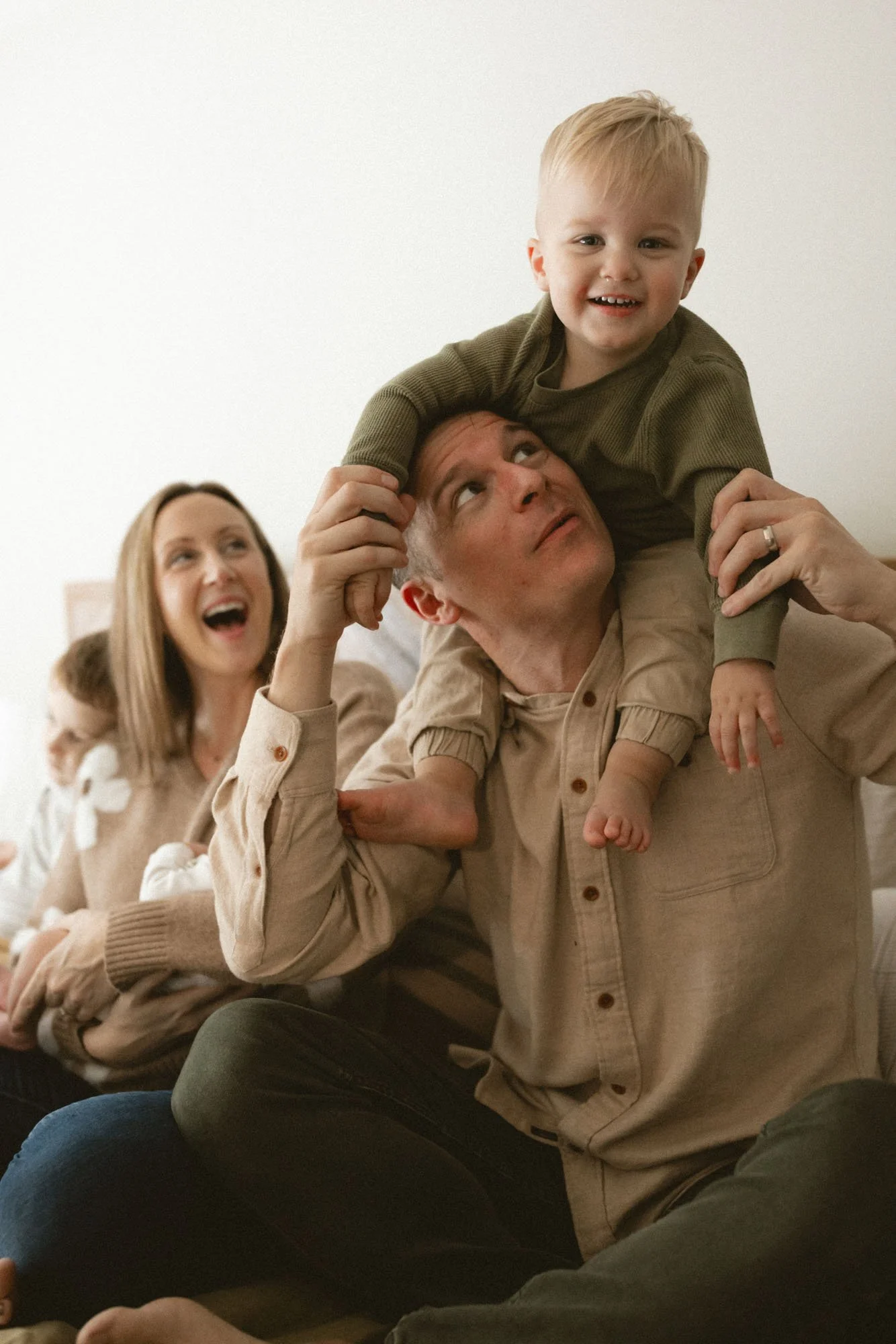 A father lifts his smiling toddler onto his shoulders while a mother holds their newborn during a cozy in-home lifestyle family photography session in West Seattle.