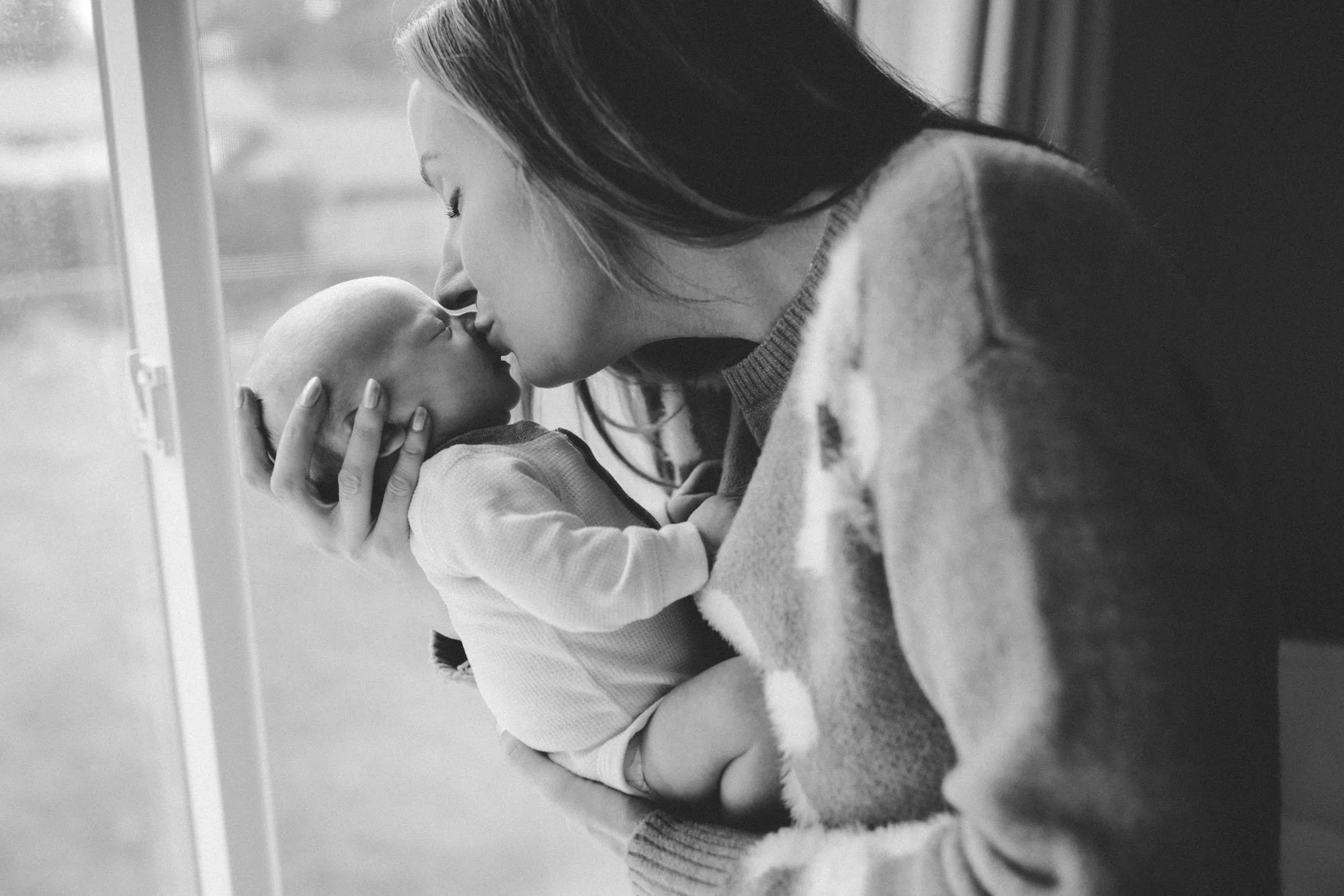 Mother gently kissing her newborn baby by a window, captured in soft natural light during an in-home lifestyle newborn session in Seattle