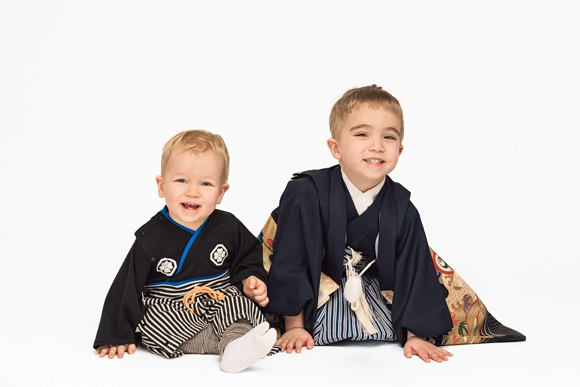 Two brothers in matching Japanese kimono outfits sitting together and smiling, simple white studio backdrop – Seattle sibling milestone and kimono photography