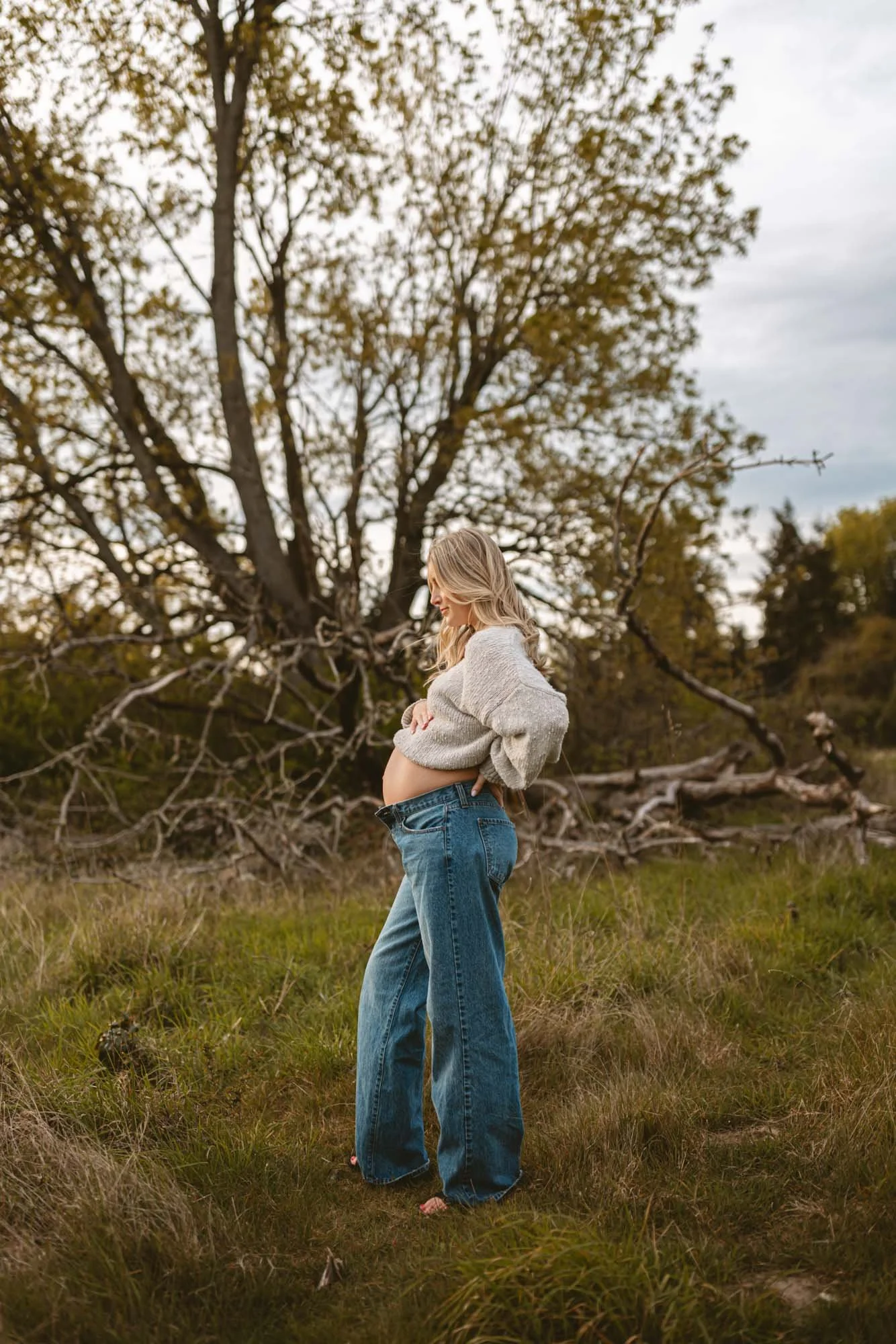 Mother-to-be standing quietly in the field at Discovery Park, gently holding her belly, surrounded by natural textures and a calm, earthy landscape.