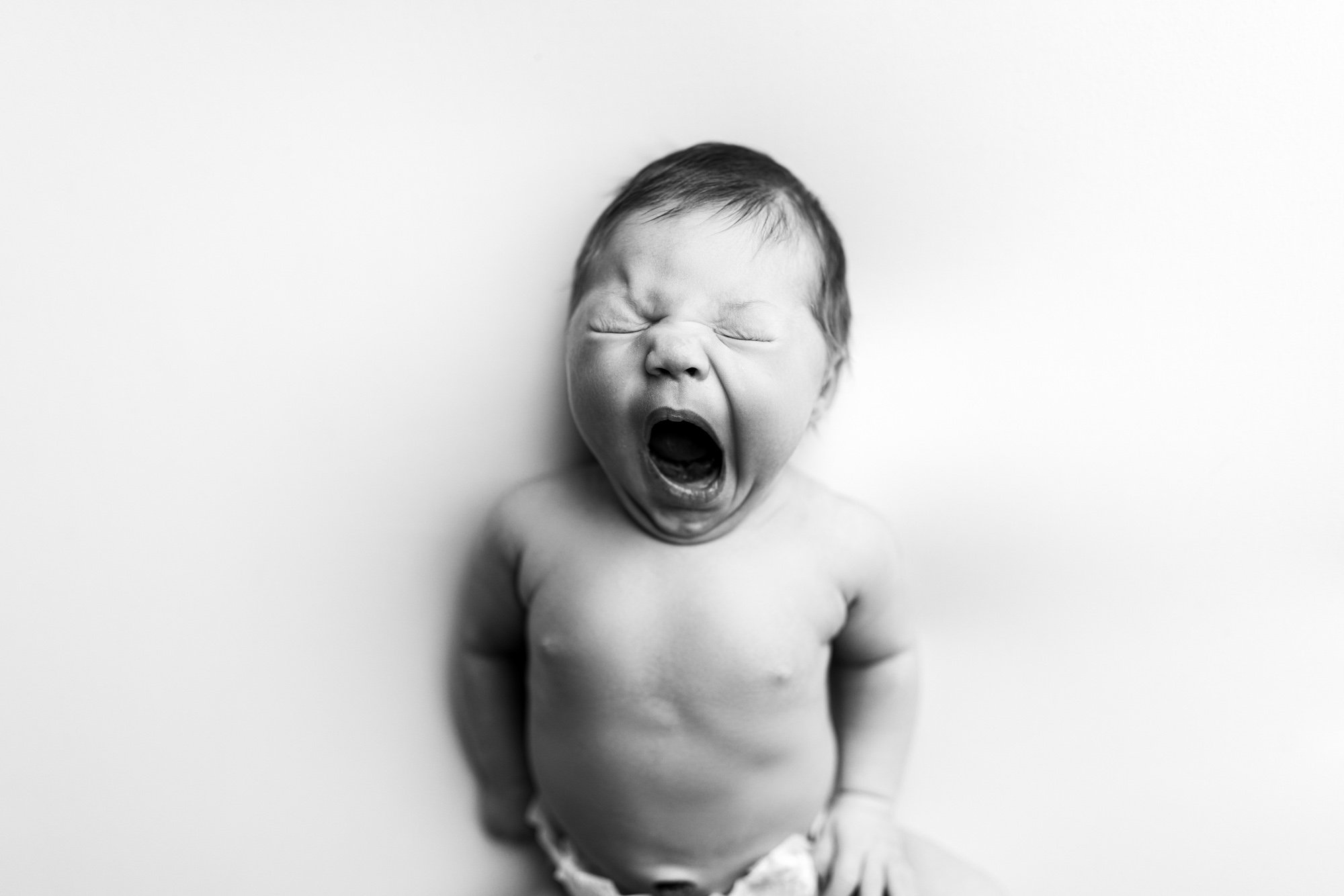 A young child, shirtless and wearing a diaper, yawning or crying with eyes closed and mouth wide open, standing against a plain white background.