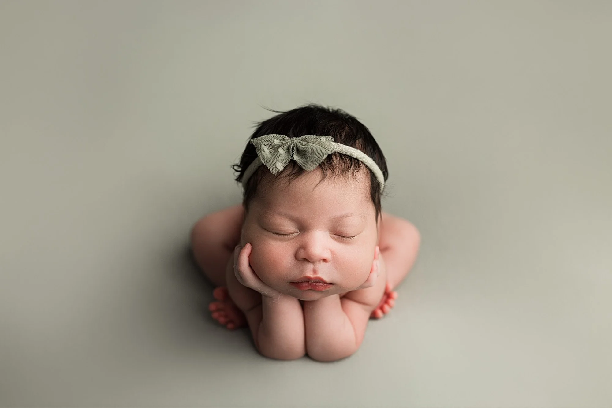 Sleeping newborn baby girl wearing a bow and posed with her hands beneath her chin