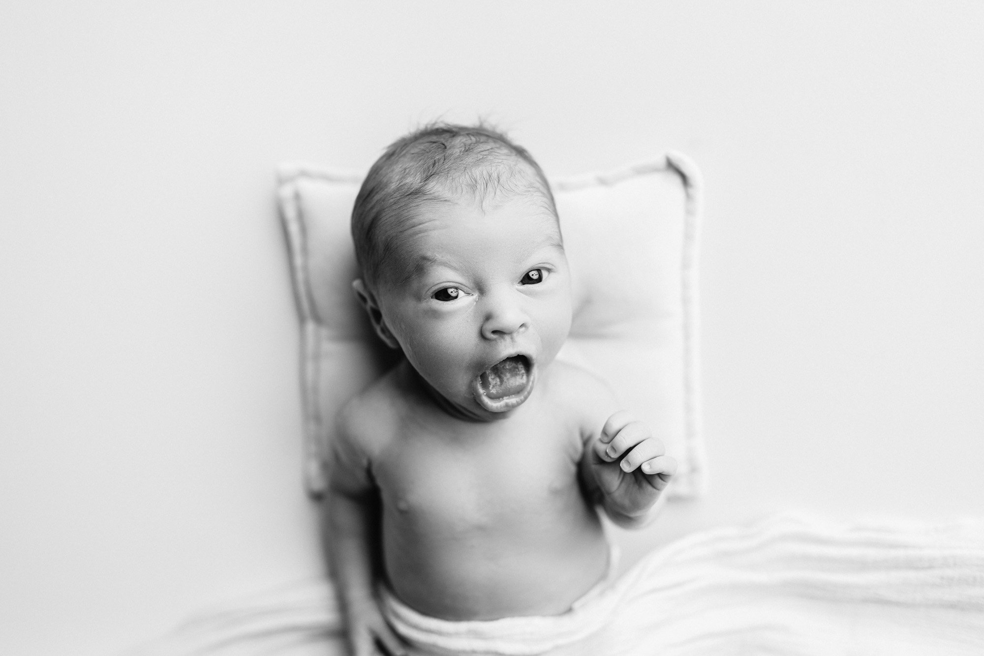 Black and white photograph of a shirtless baby lying on a pillow, with its mouth open and eyes wide, looking upwards.