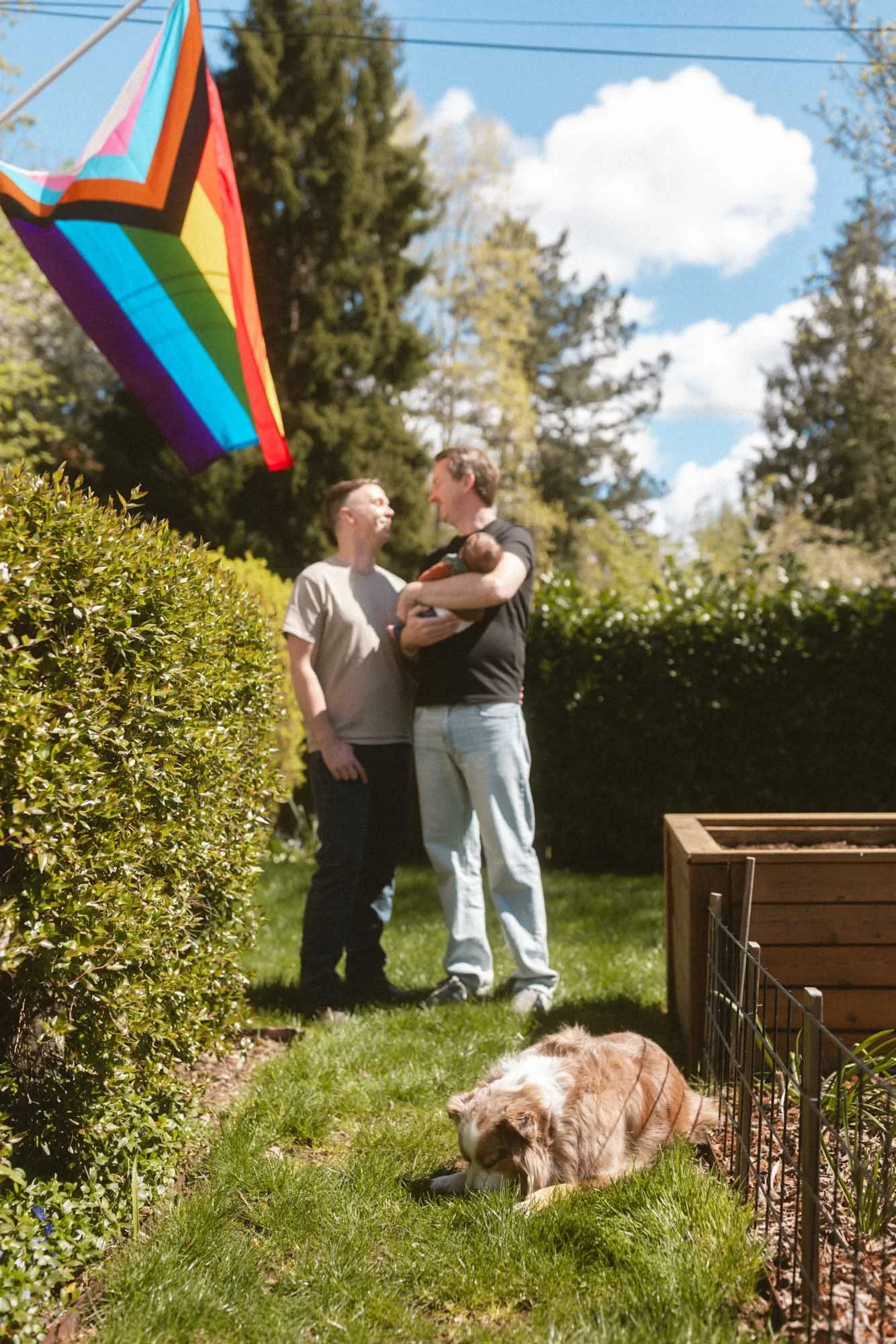 A joyful outdoor portrait of two dads holding their newborn beneath a rainbow flag, celebrating family, identity, and love in a bright natural setting.