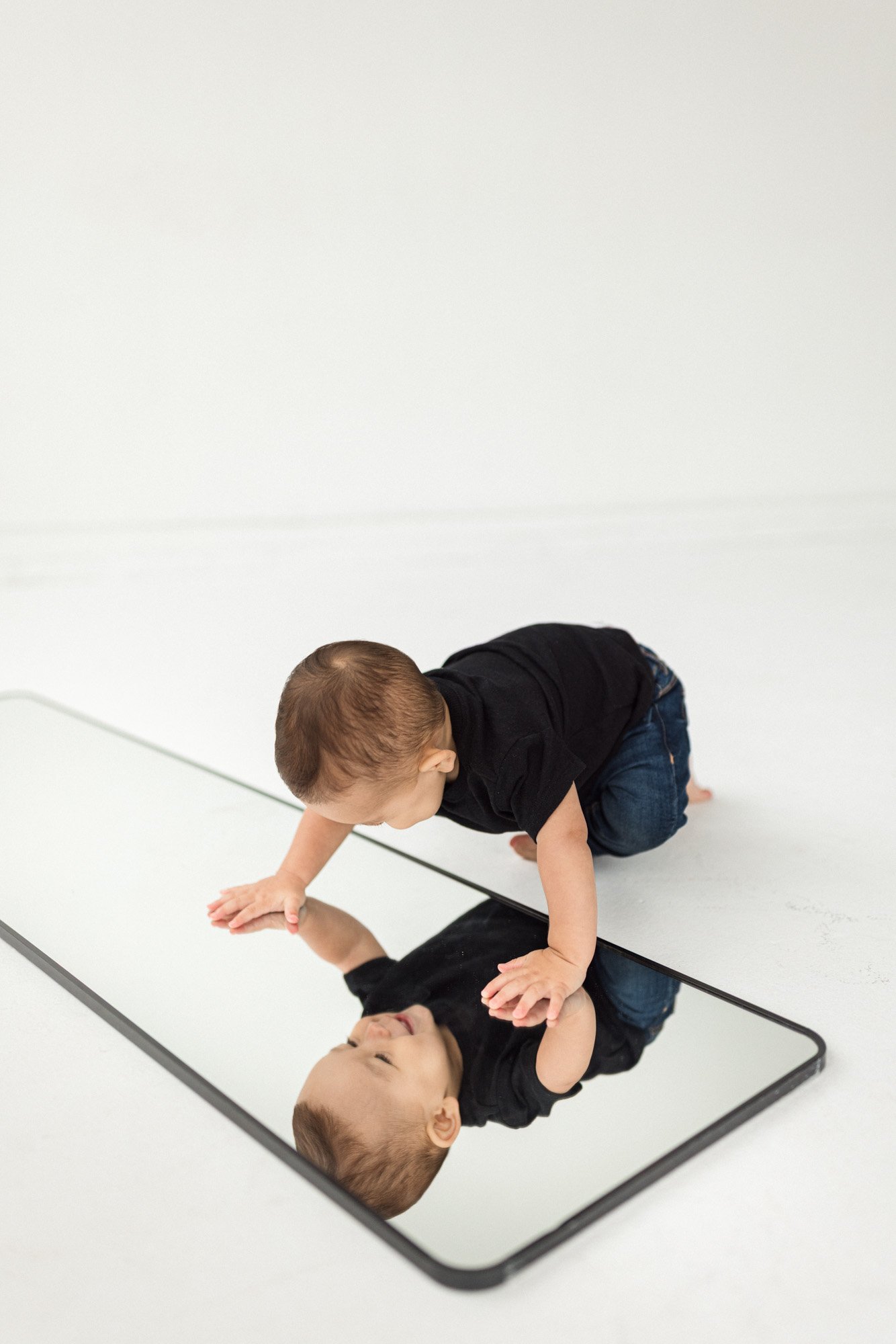 Curious baby exploring his reflection in a mirror during a minimalist studio milestone session, photographed by a Seattle baby photographer. Clean white backdrop, natural tones, and candid movement capture early developmental milestones in a modern l