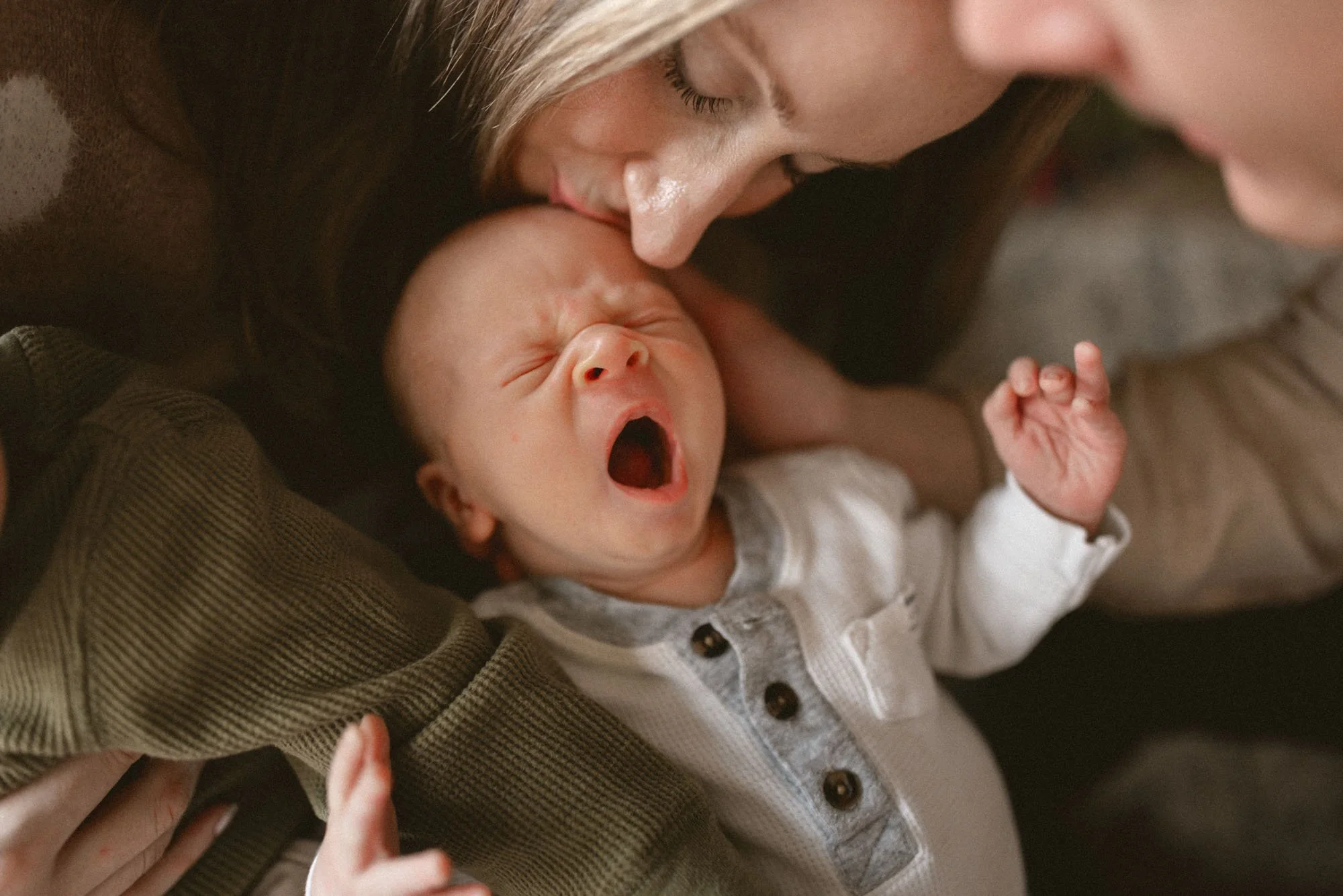 A mother gently kisses her yawning newborn during a cozy in-home newborn photography session in Seattle.