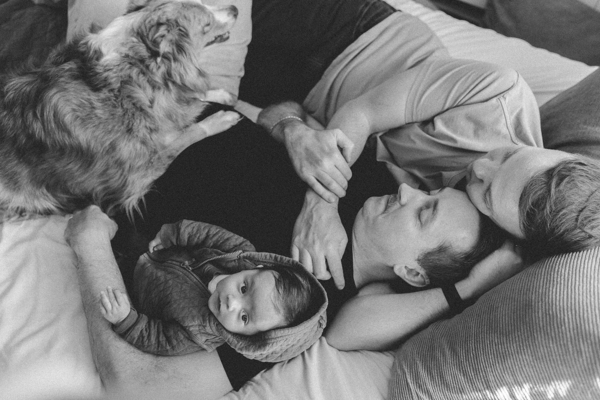Black and white portrait of gay dads cuddling their newborn baby with their dog during an in-home session.