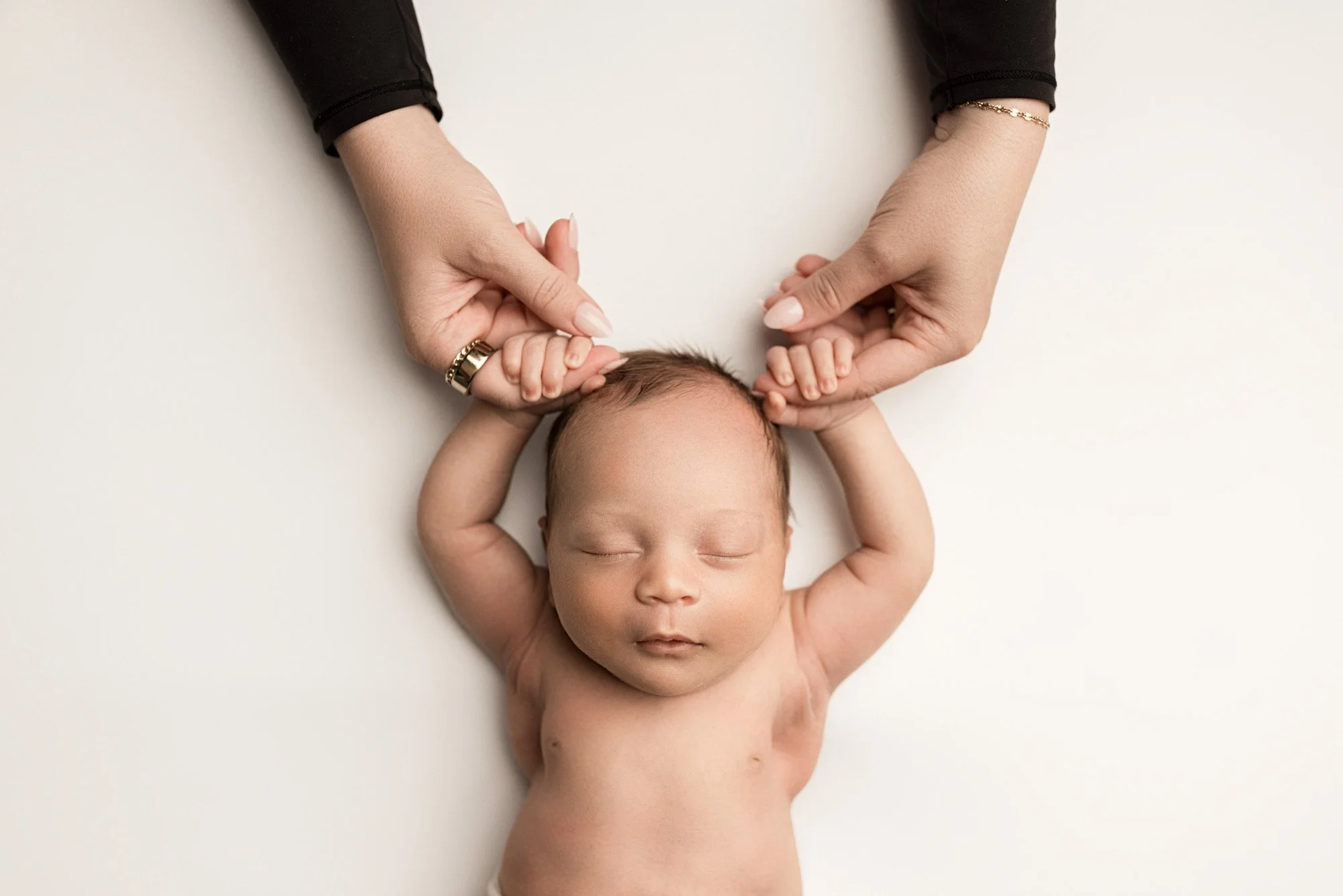 Mother gently holding her sleeping newborn baby’s hands on a soft white backdrop