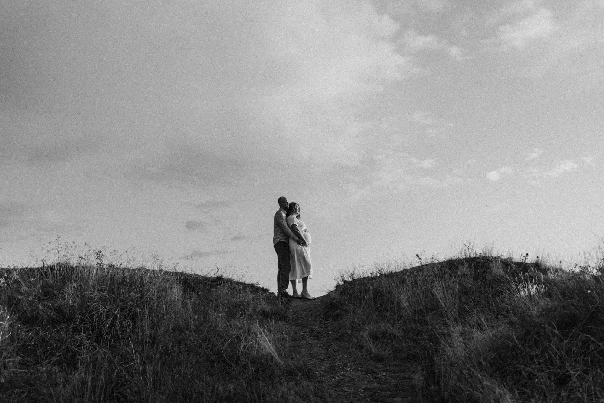 Expecting couple standing together on a hill at sunset, embracing with baby bump visible, black and white maternity portrait in Seattle