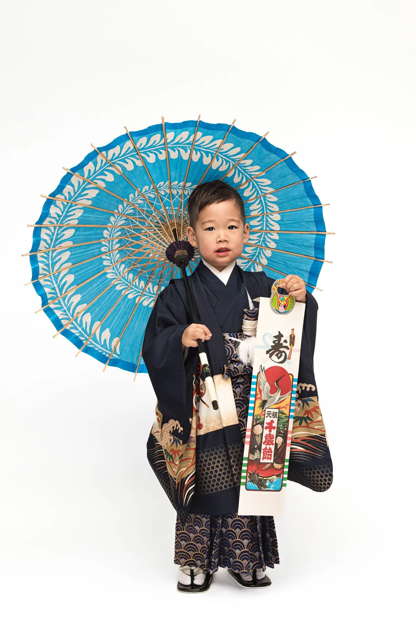 Young boy in navy traditional kimono holding a ceremonial bag and standing with a blue umbrella during a Shichigosan portrait session in a Seattle studio