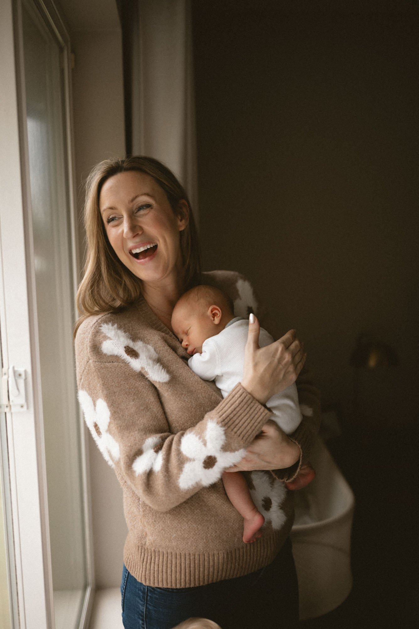 A mother smiles while holding her sleeping newborn in soft window light during an in-home newborn photography session in Lake Tapps, WA.