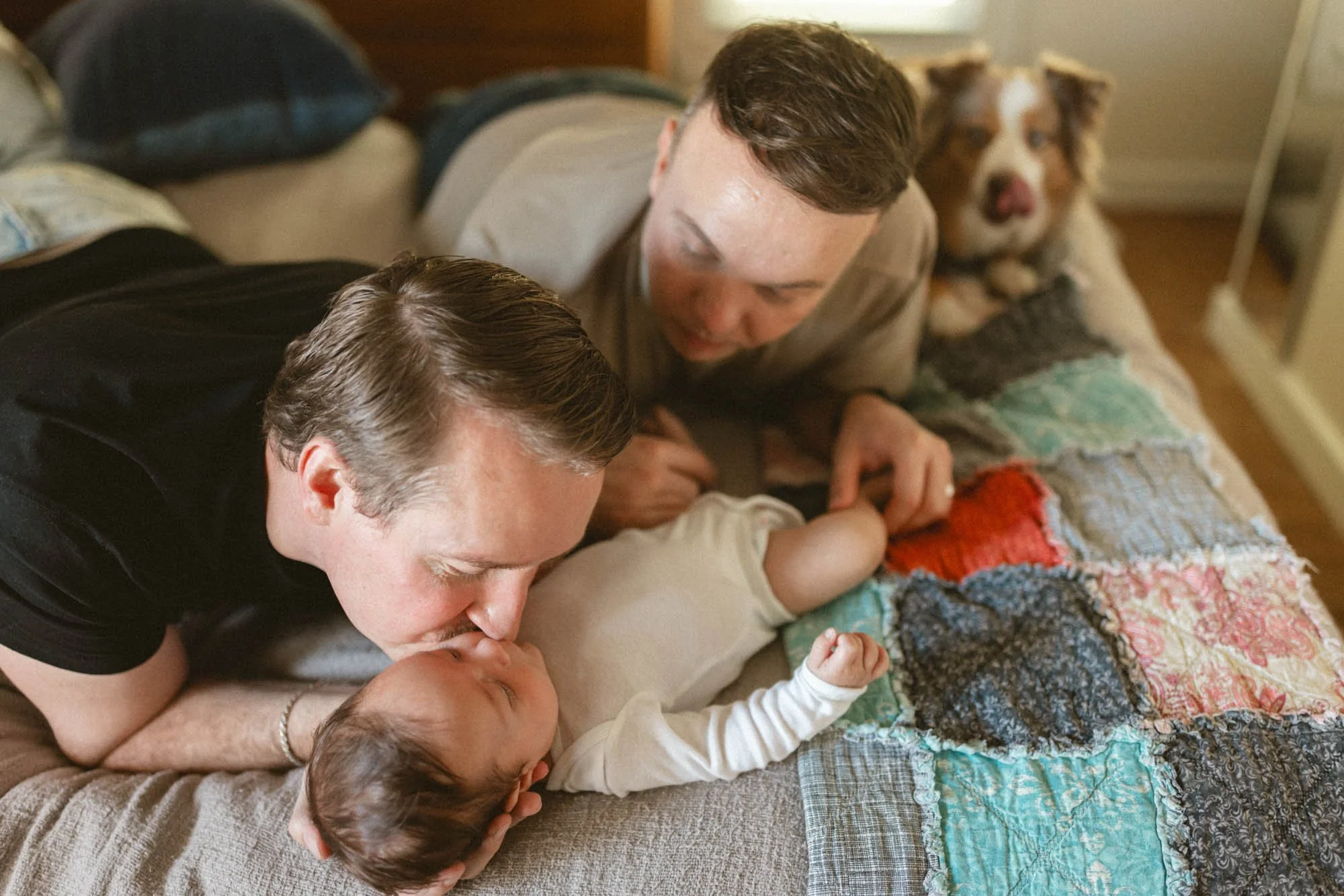 An intimate in-home newborn session featuring two dads gently kissing their baby on a cozy couch, documenting real connection, love, and the early days of parenthood.