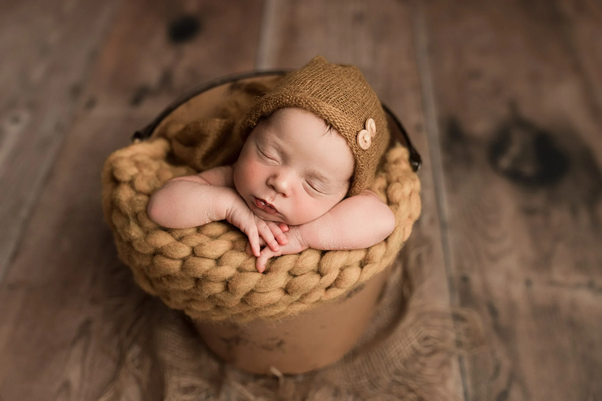 Newborn baby boy sleeping in a rustic bucket setup with a knitted bonnet during a cozy studio newborn photography session near Seattle, WA.