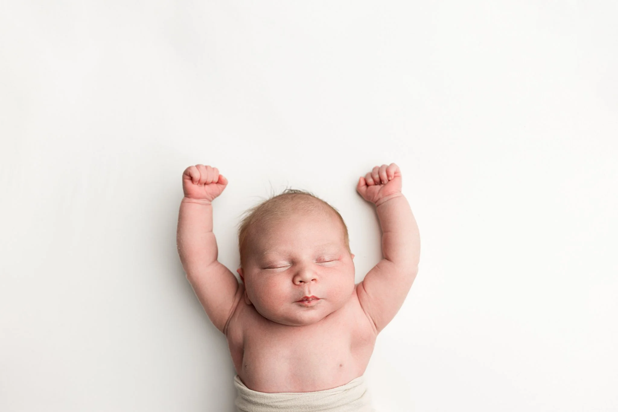 Newborn baby with arms raised sleeping against a clean white backdrop during a minimalist studio newborn photography session in Seattle.