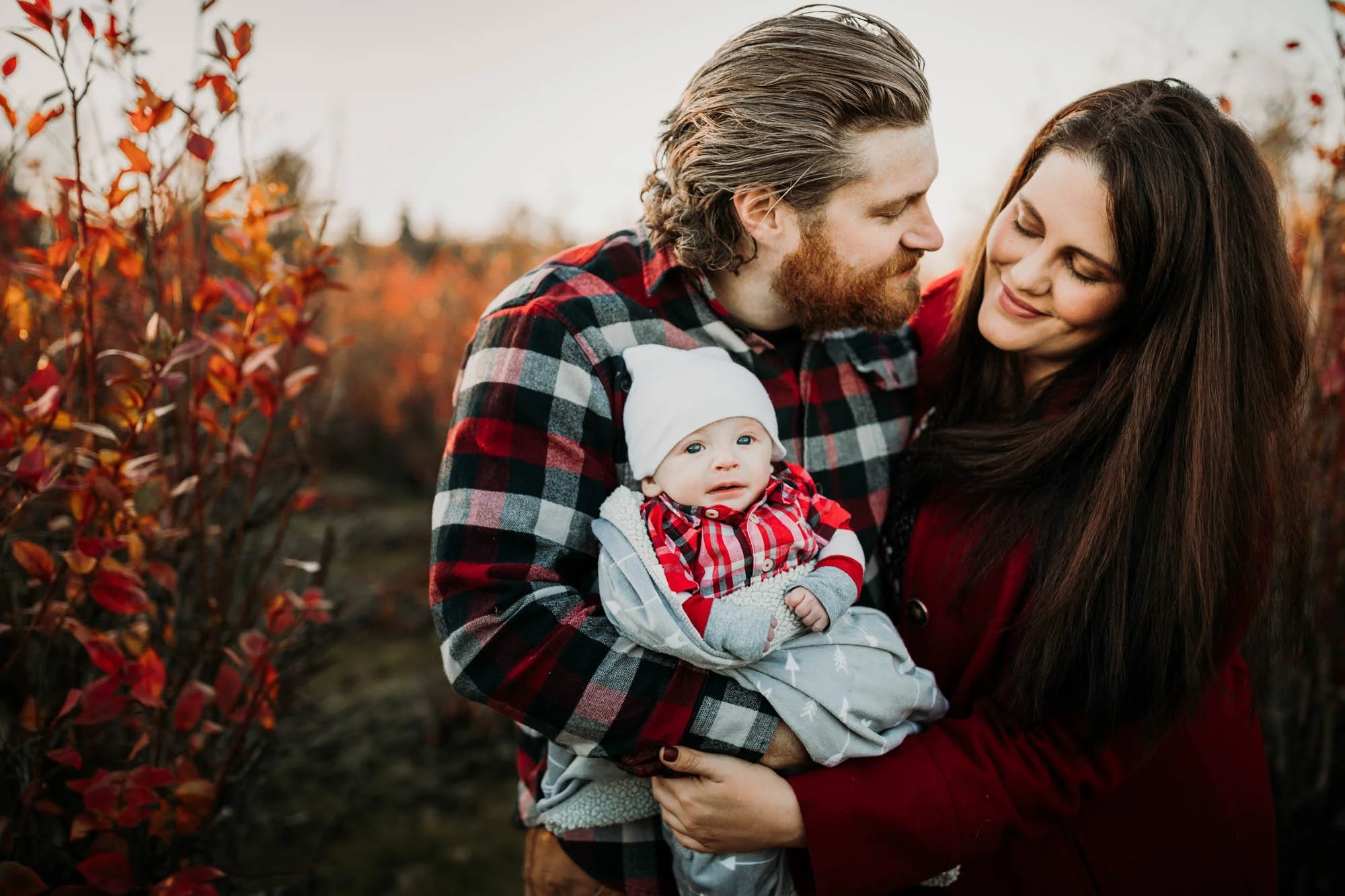 A family of three, a man, a woman, and a baby, outdoors during fall. The man has a beard and is wearing a red and black plaid shirt, holding the baby. The woman has long dark hair and is wearing a red coat, leaning towards the man. The baby is dressed in a red plaid shirt, a white hat, and a blanket, looking at the camera with blue eyes. Fall foliage with red and orange leaves surrounds them.