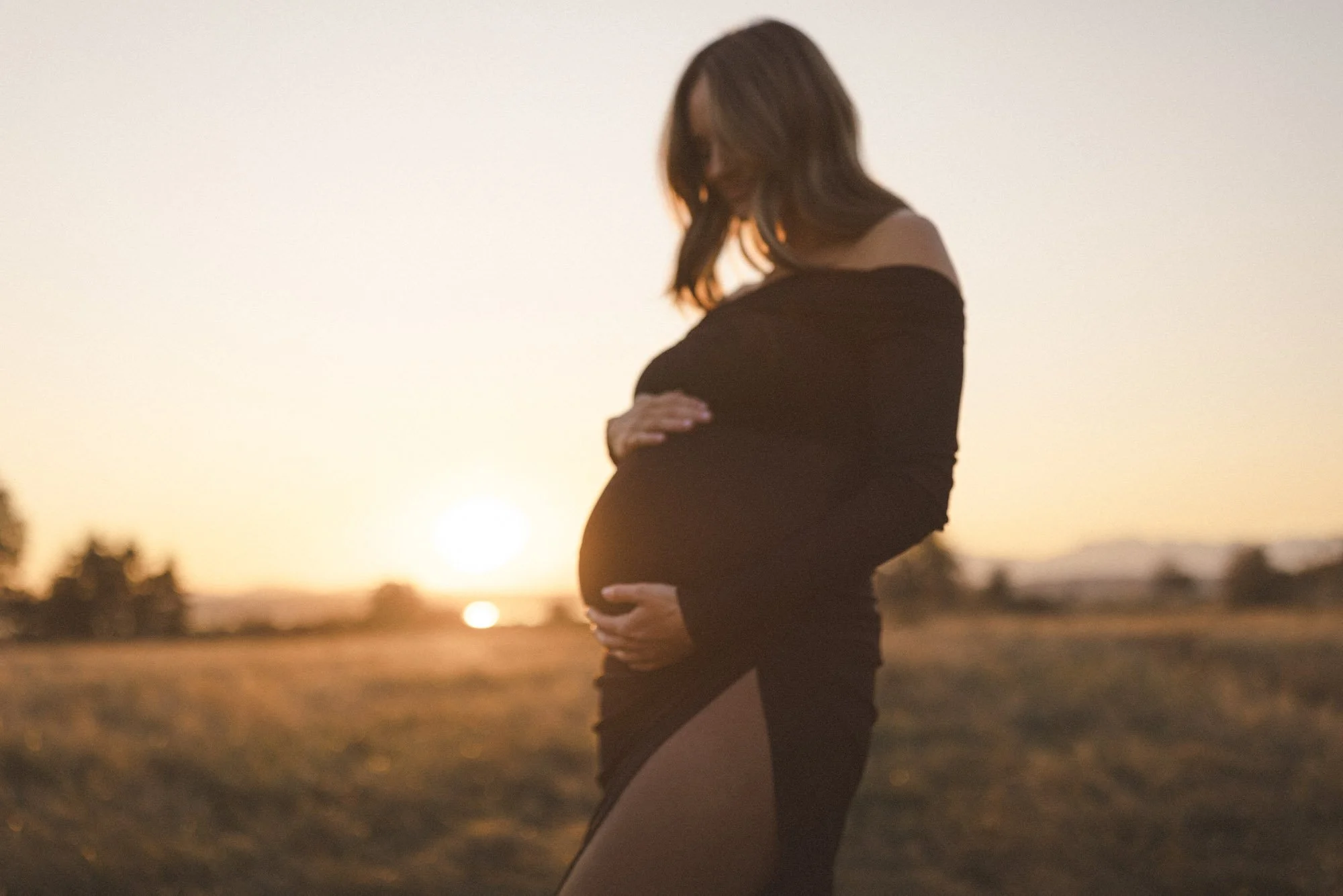 Seattle maternity photo of expecting mother in black dress holding baby bump at sunset in a field, golden hour pregnancy portrait