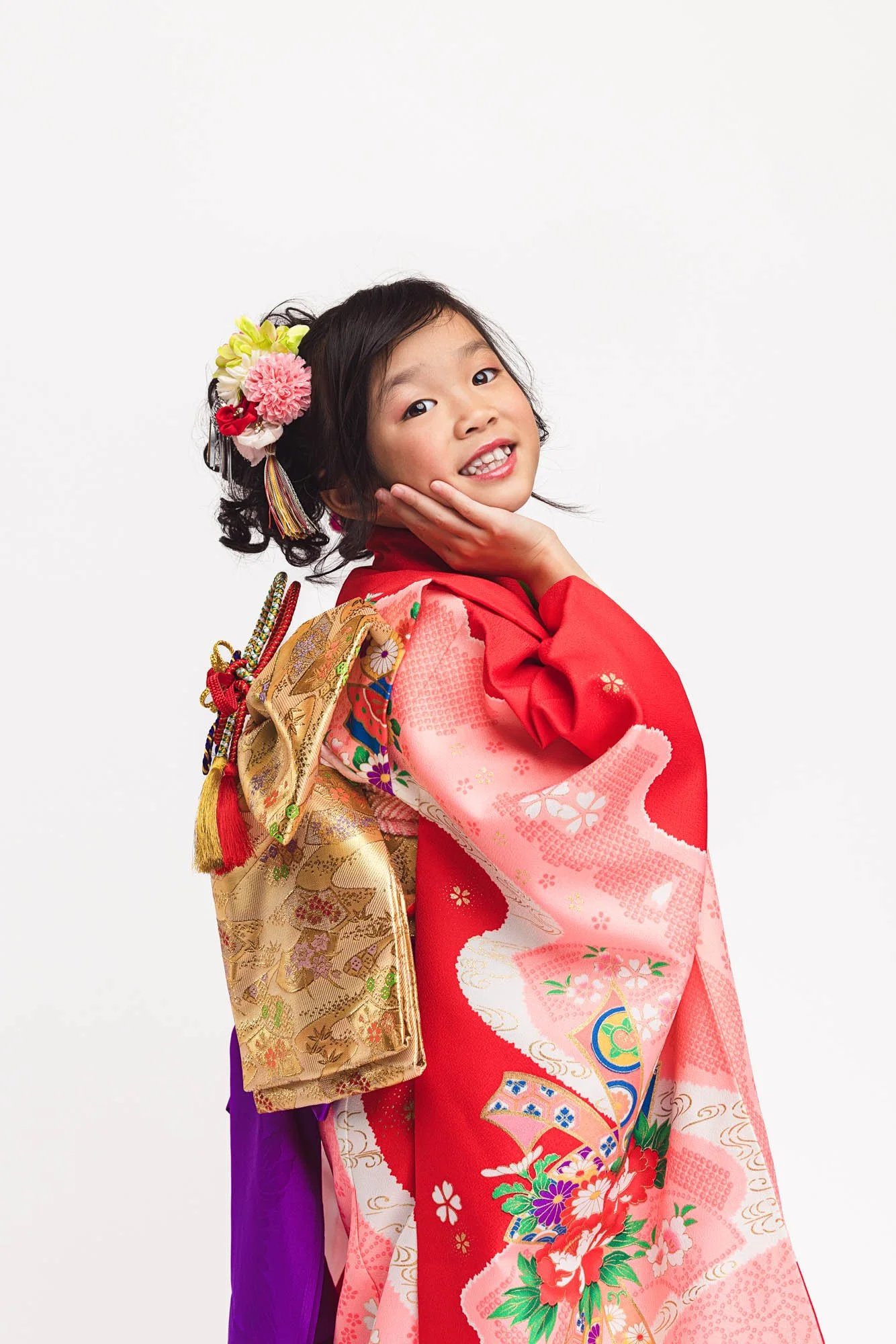Young girl in red and pink traditional kimono smiling over her shoulder during a Shichigosan portrait session in a Seattle studio