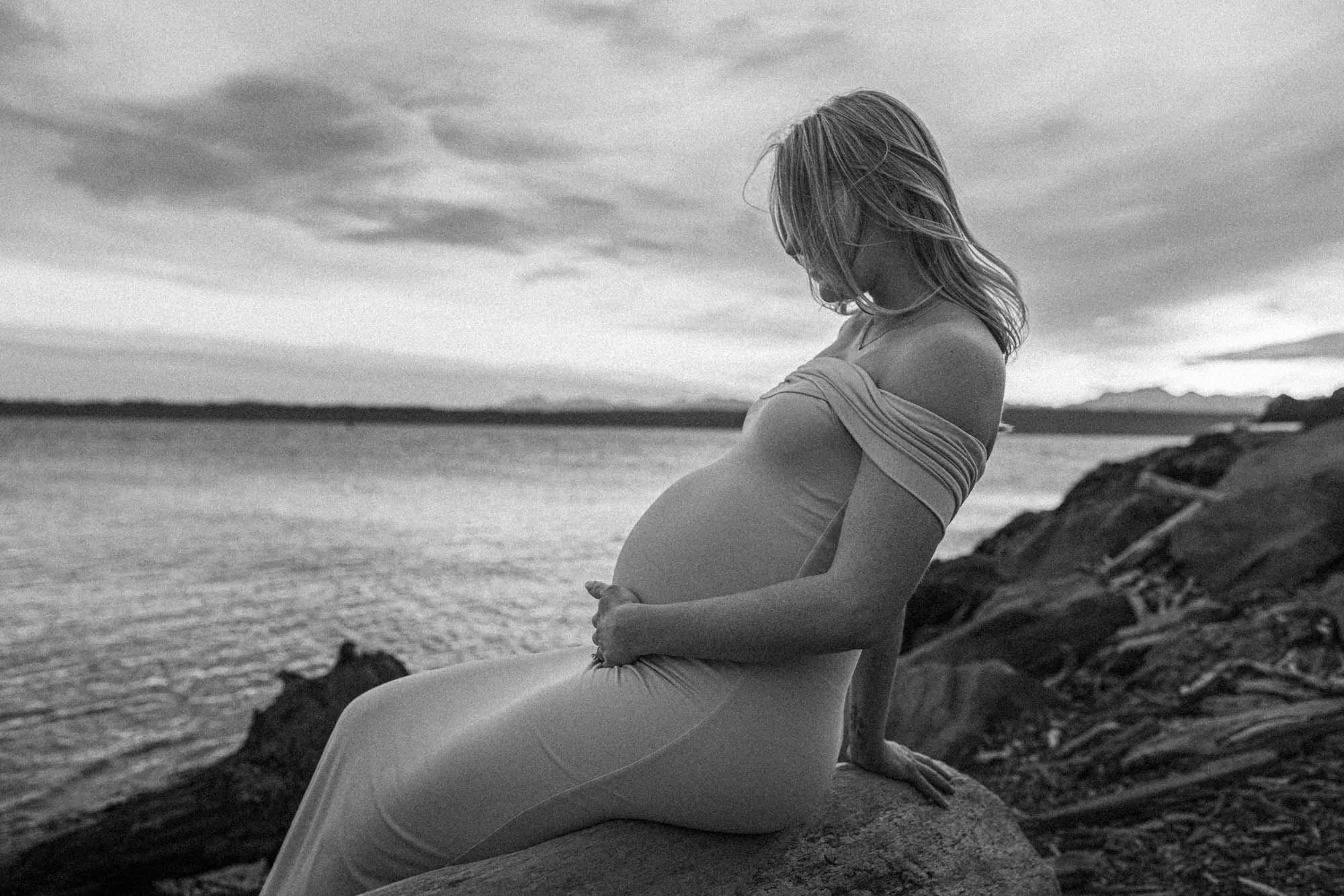 Pregnant mother sitting by the shoreline at Discovery Park in Seattle, gently holding her belly with a calm, reflective mood against the coastal backdrop.